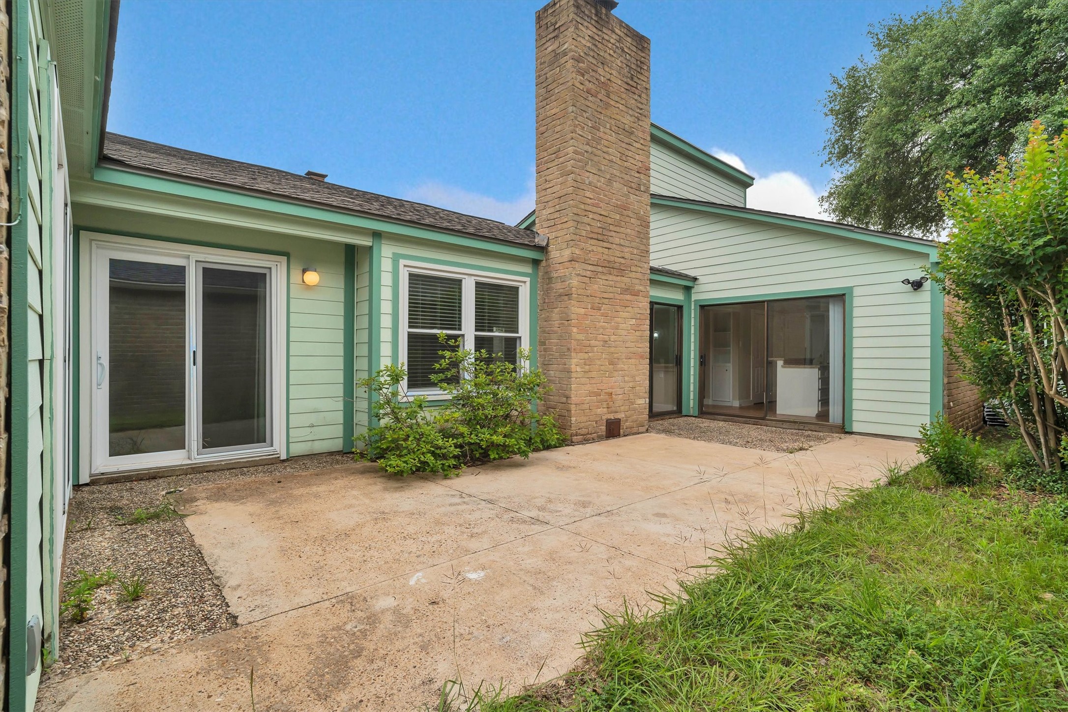 2163 Paso Rello Drive Houston, TX 77077 - Photo 3 of 45 cozy backyard with a concrete patio, sliding glass doors, light green siding, and a central brick chimney. It's bordered by a small garden and has potential for outdoor relaxation or entertaining.