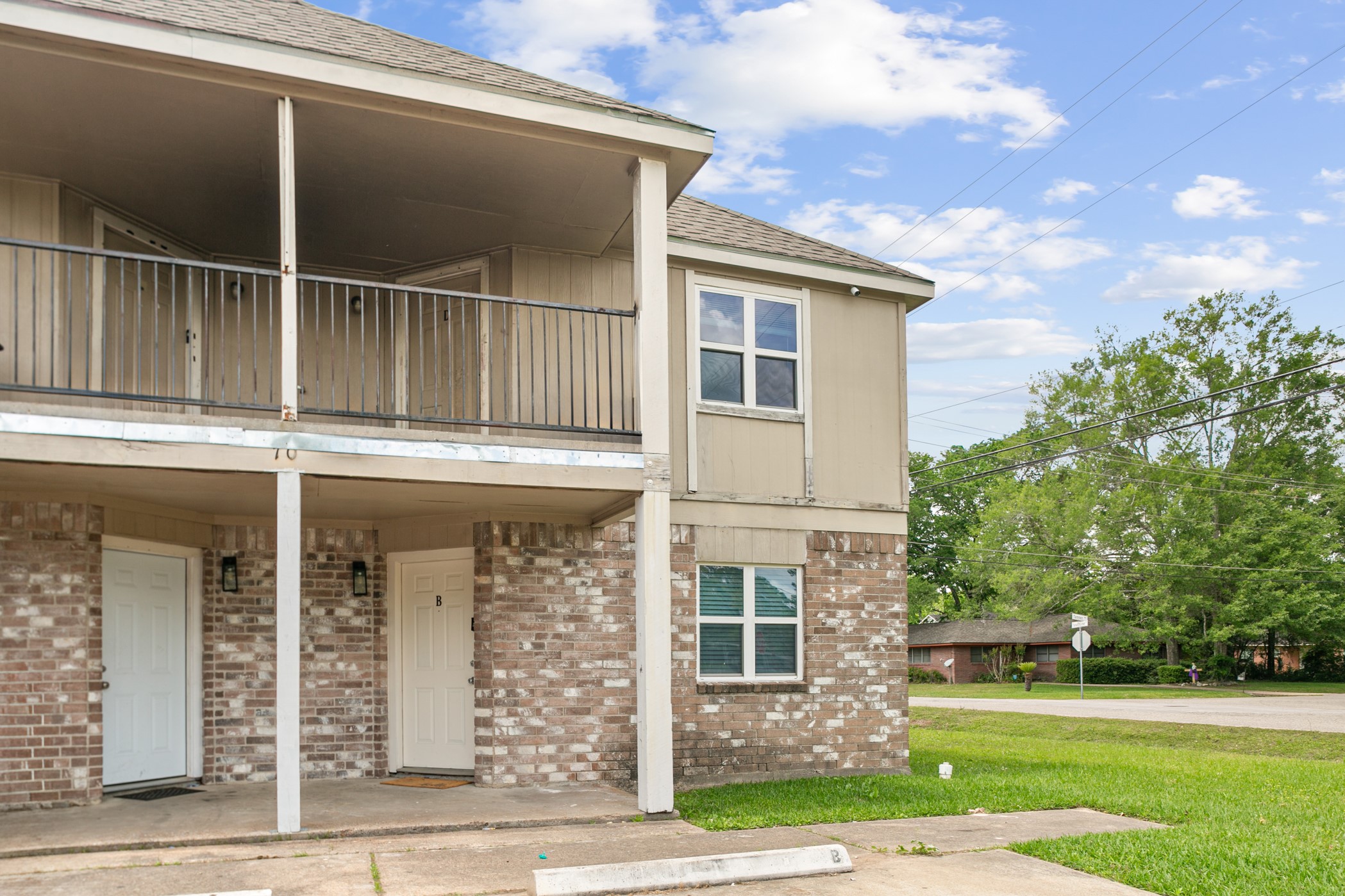 700 Lyle W Avenue, Unit D Cleveland, TX 77327 - Photo 16 of 16 This photo shows a two-story townhouse with a brick and siding exterior. It features a covered upper balcony and two front entrances, situated on a corner lot with a grassy lawn. The area appears residential and quiet, with trees and a clear sky in the background.