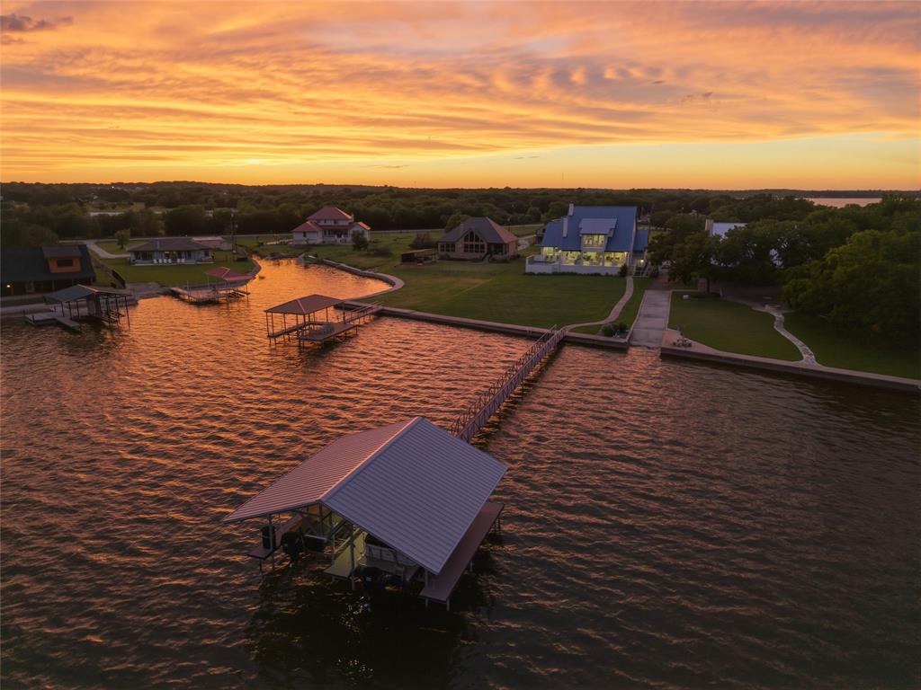 6625 Sandy Beach Road Azle, TX 76020 - Photo 3 of 40 Aerial view showcases the waterfront positioning of the property with extended shoreline frontage and private dock access. Rear elevation includes expansive windows, covered outdoor living areas, and landscaped grounds extending toward the water. Concrete walkways, mature trees, and surrounding waterfront properties are visible throughout the aerial perspective.