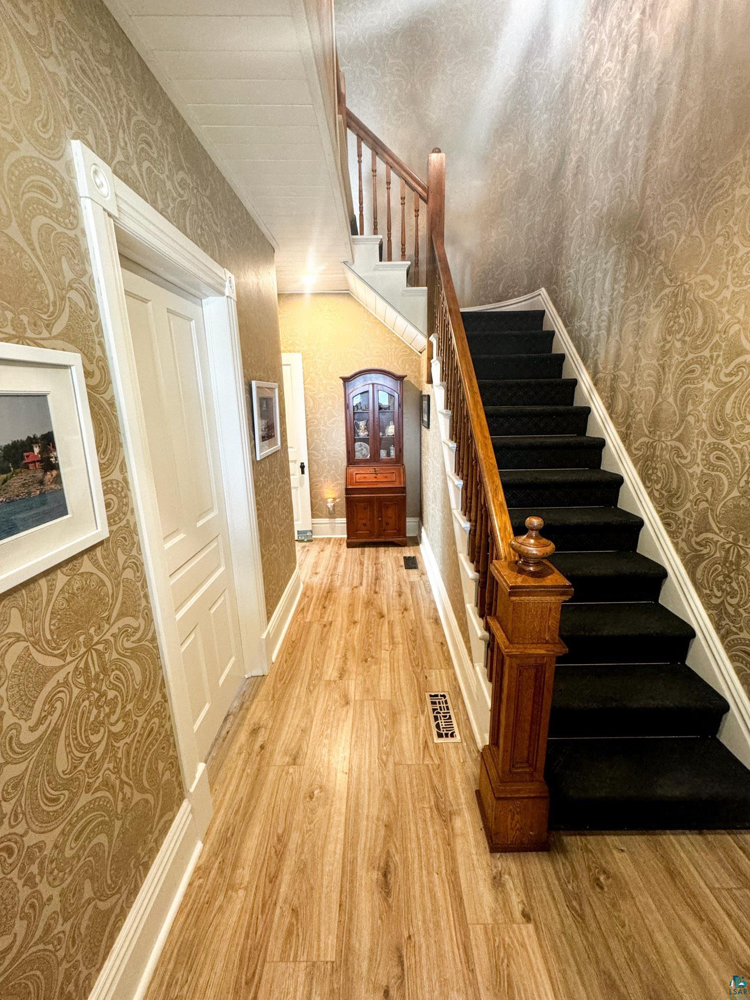 303 East 5th Street Washburn, WI 54891 - Photo 22 of 34 Hallway featuring wood-finish flooring, patterned wall coverings, white trim, and a staircase with a wooden banister and carpeted treads