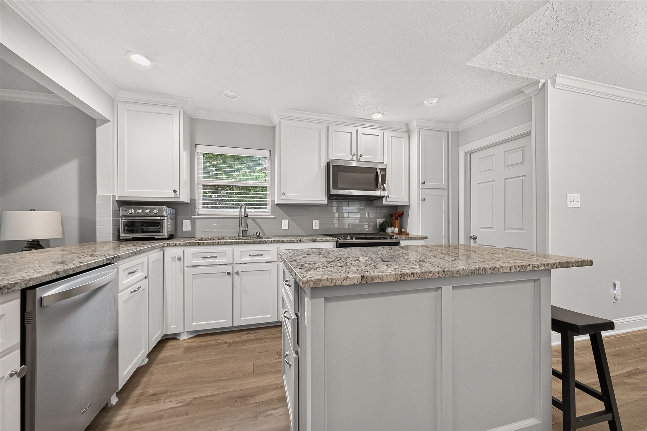 24719 Wilderness Road Spring, TX 77380 - Photo 17 of 42 This kitchen features modern white cabinetry, granite countertops, and stainless steel appliances, including a microwave and dishwasher. A large island offers additional prep space and seating, complemented by ceramic tile flooring and recessed lighting for a bright, inviting atmosphere.