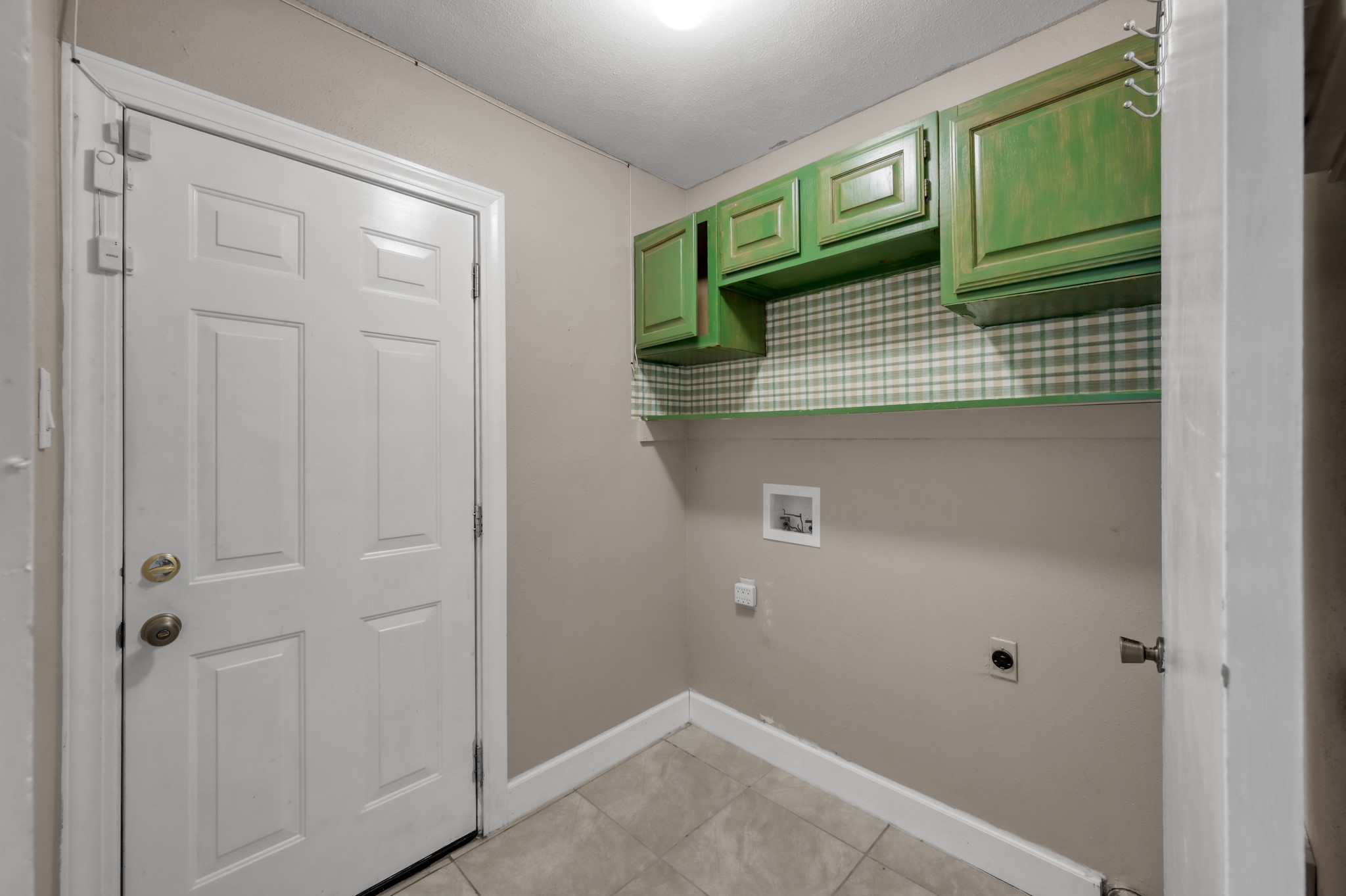 12015 Fallbrook Drive Houston, TX 77065 - Photo 14 of 29 This photo shows a small laundry room with a tiled floor, white entry door, and green cabinets above washer/dryer hookups. The walls are neutral with a plaid accent.