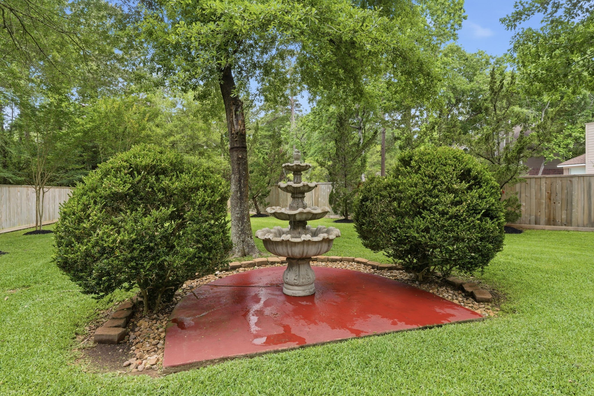 11 Dovewing Place Spring, TX 77382 - Photo 29 of 32 Backyard features a charming, tiered fountain on a red concrete pad, surrounded by lush greenery and manicured shrubs, offering a peaceful, private retreat.