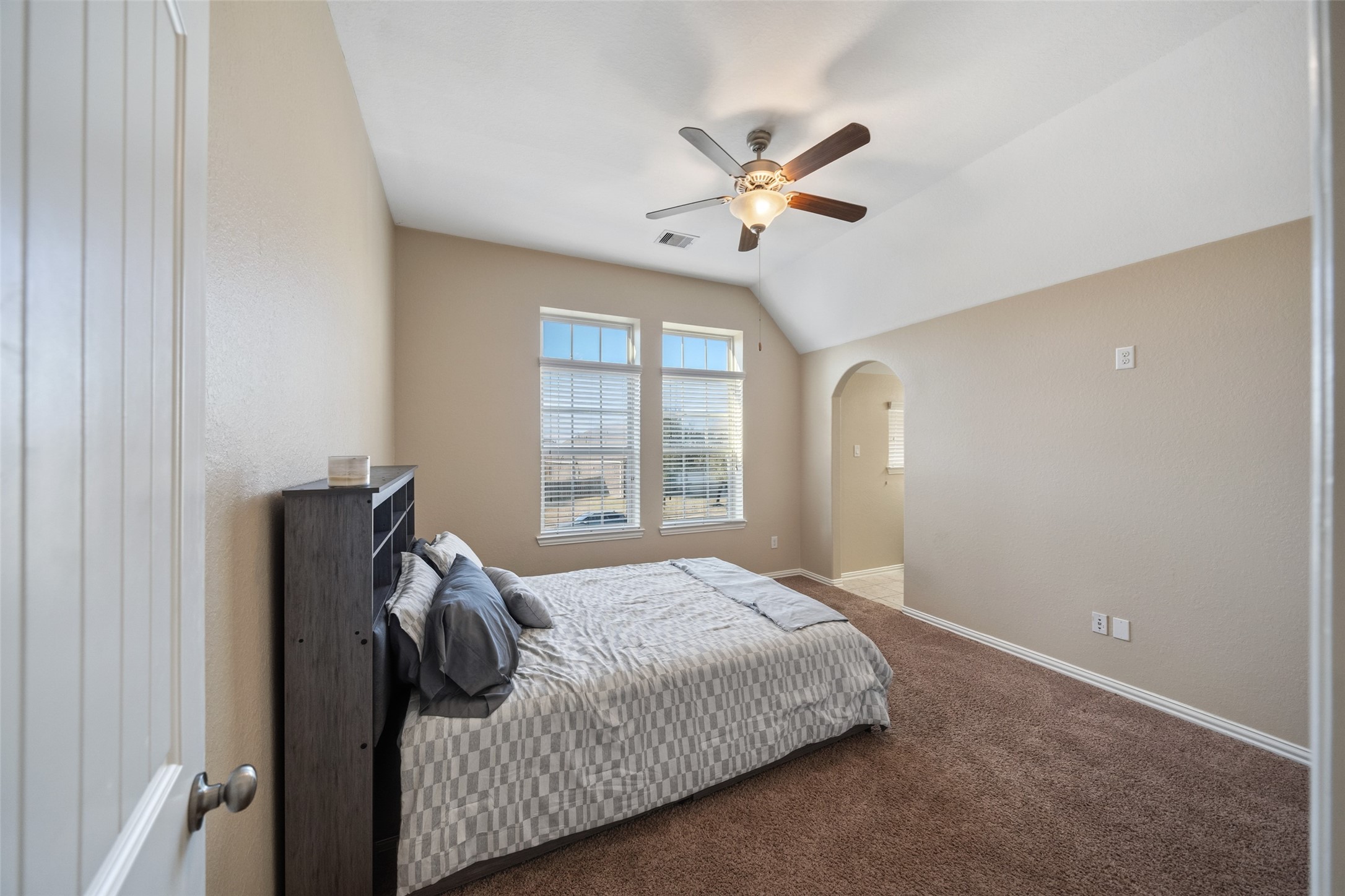 4747 Abercorn Street Rosharon, TX 77583 - Photo 26 of 46 This bedroom features neutral walls and carpet, a ceiling fan, and ample natural light from the large windows. It includes a cozy bed and leads into an adjacent area through an arched doorway.