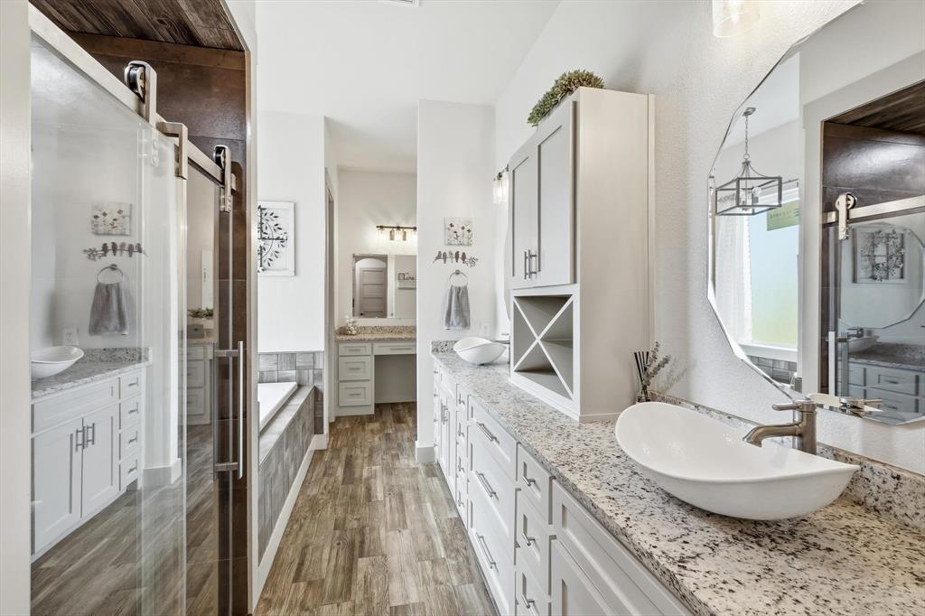 6020 Honeysuckle Road Midlothian, TX 76065 - Photo 20 of 39 Dual vessel sinks set on a light granite-finish countertop, expansive vanity cabinetry, wood-look flooring, and a large soaking tub with a tiled surround