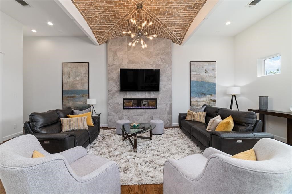 710 Delano Street, Unit A Houston, TX 77003 - Photo 3 of 32 The living room features a vaulted brick ceiling, modern chandelier, and a sleek stone fireplace with a mounted TV. It includes stylish leather sofas, cozy armchairs, and a plush area rug, creating an inviting space for relaxation and entertainment.