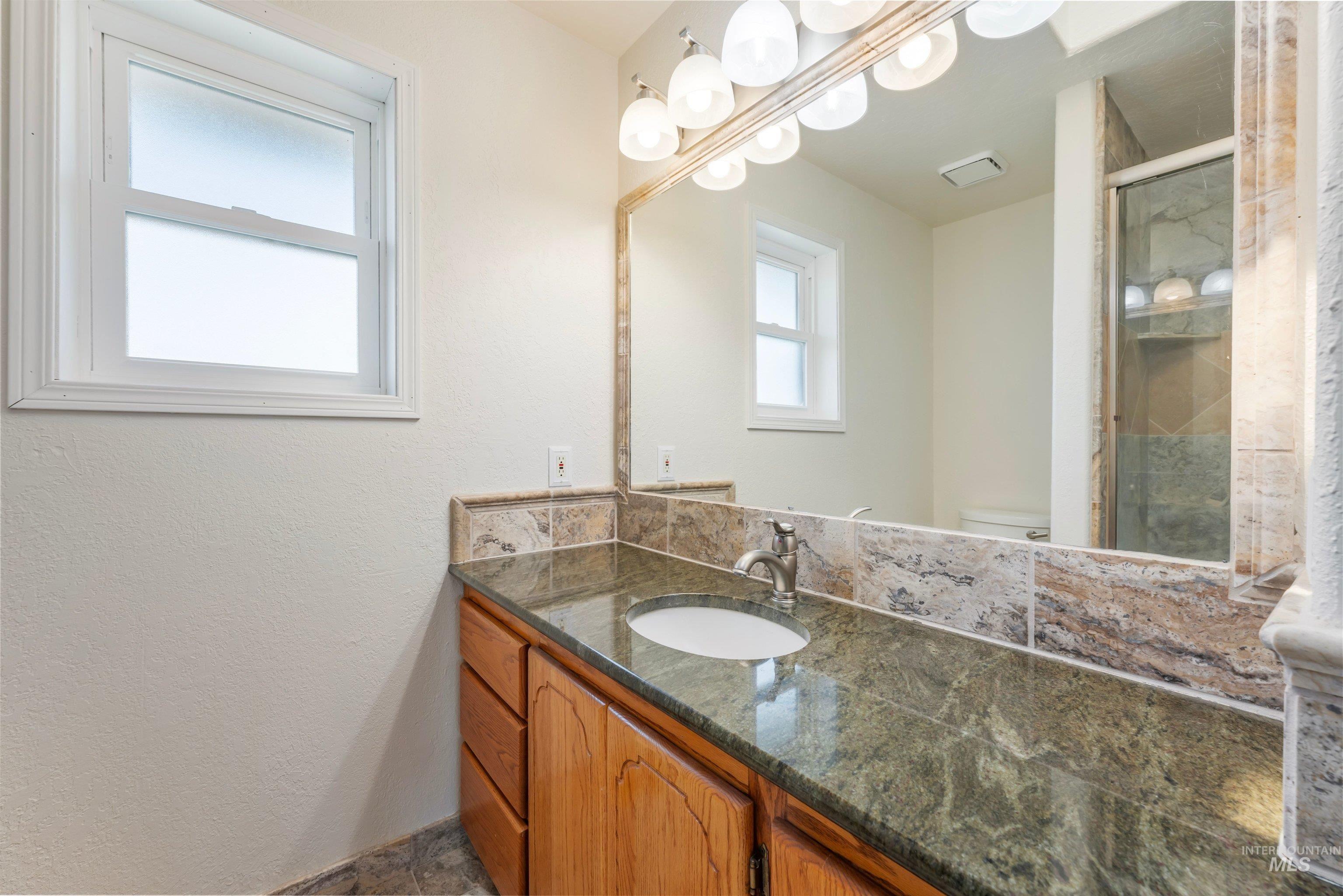 7657 Track Road Nampa, ID 83686 - Photo 29 of 49 Bathroom vanity featuring a green stone countertop with an integrated sink, wood cabinetry, and a full-width mirror with overhead lighting