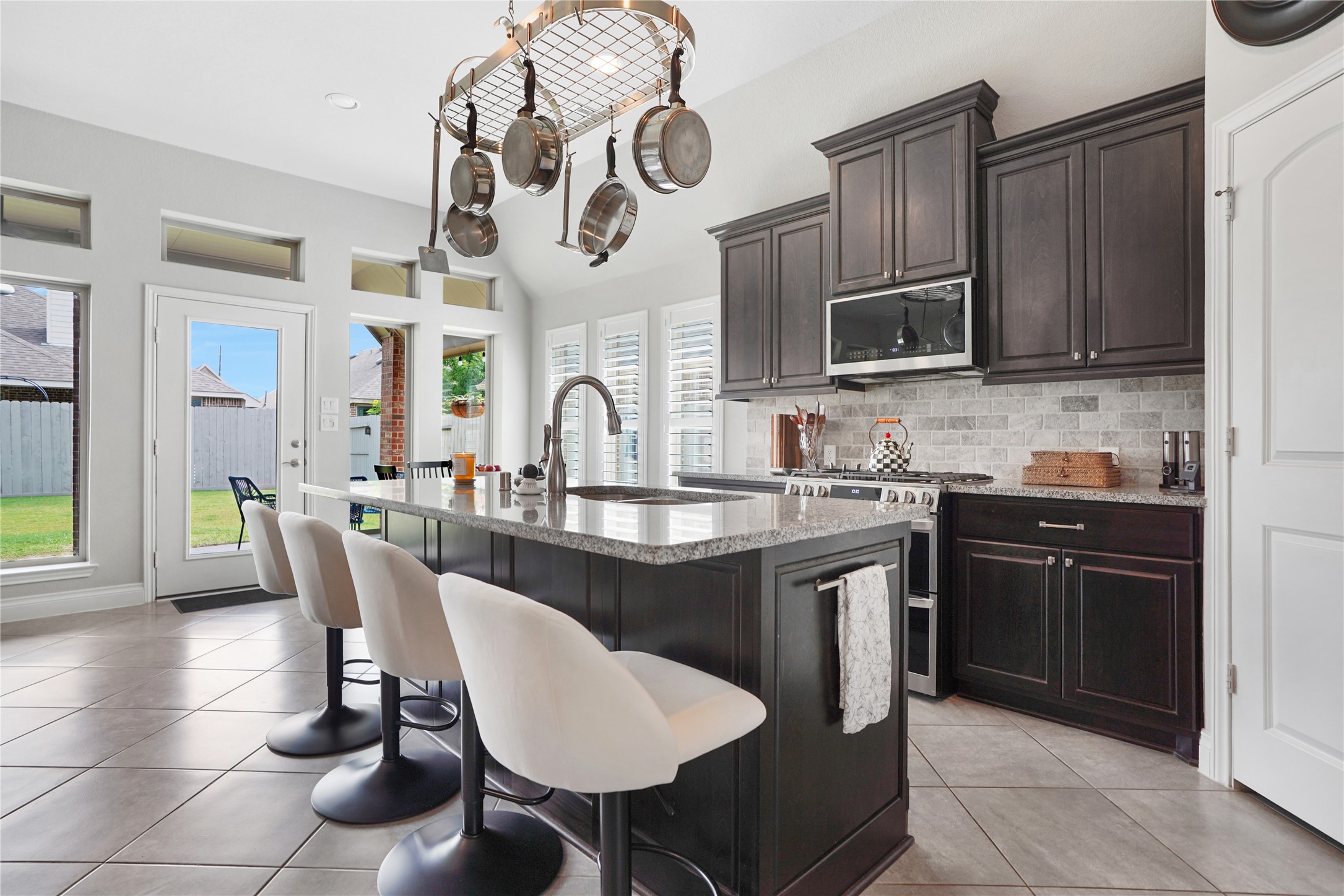 2345 Redwood Ridge Trail Manvel, TX 77578 - Photo 12 of 39 This modern kitchen features dark cabinetry, a central island with seating, and a pot rack. It is brightened by multiple windows and a glass door leading to a backyard, creating a spacious and inviting atmosphere.