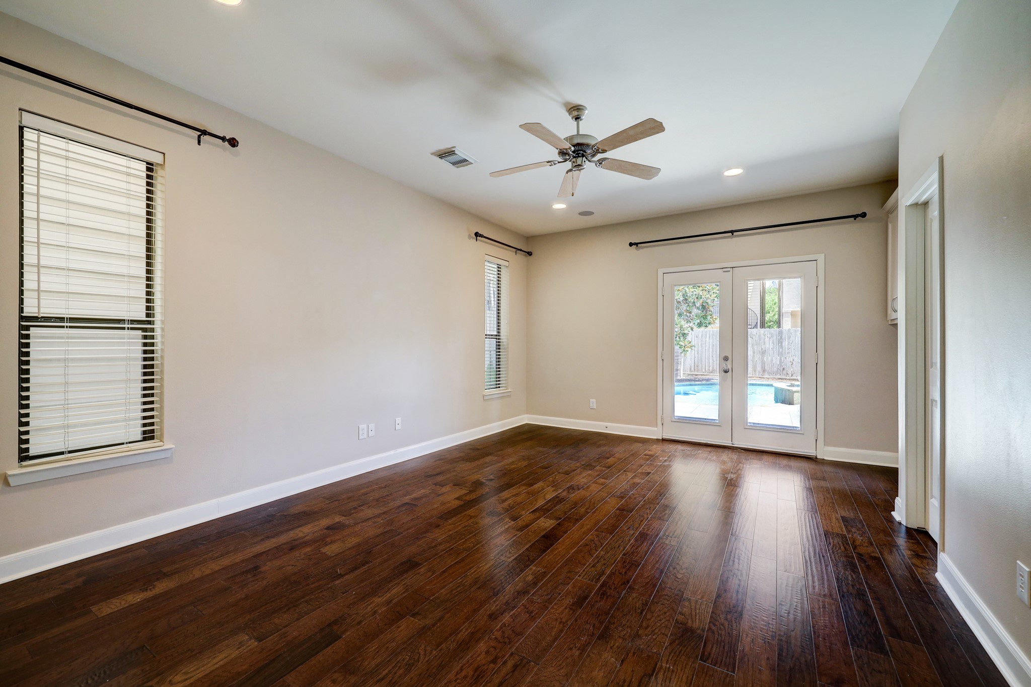 5202 Rose Street, Unit B Houston, TX 77007 - Photo 26 of 40 This spacious room features rich hardwood floors, neutral walls, and ample natural light through two windows and French doors. The doors lead to a backyard with a pool, adding a relaxing outdoor element. A ceiling fan provides comfort and style.