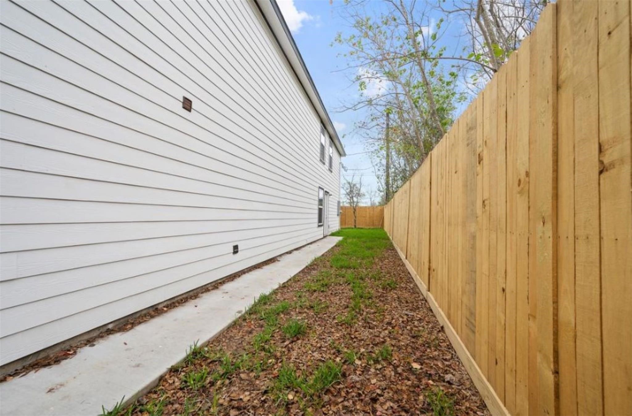 8120 Laura Koppe Road, Unit B Houston, TX 77028 - Photo 23 of 25 This photo shows a narrow side yard of a house with white siding, bordered by a tall wooden fence. The area features a concrete path and some grass, providing a private outdoor space.