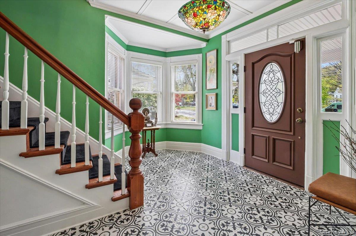 46 Bedford Road Katonah, NY 10536 - Photo 4 of 34 Entry foyer featuring patterned tile flooring, a dark wood entry door with an oval decorative glass insert, a wood staircase with white balusters, a bay window with blinds, and a stained-glass style ceiling light fixture