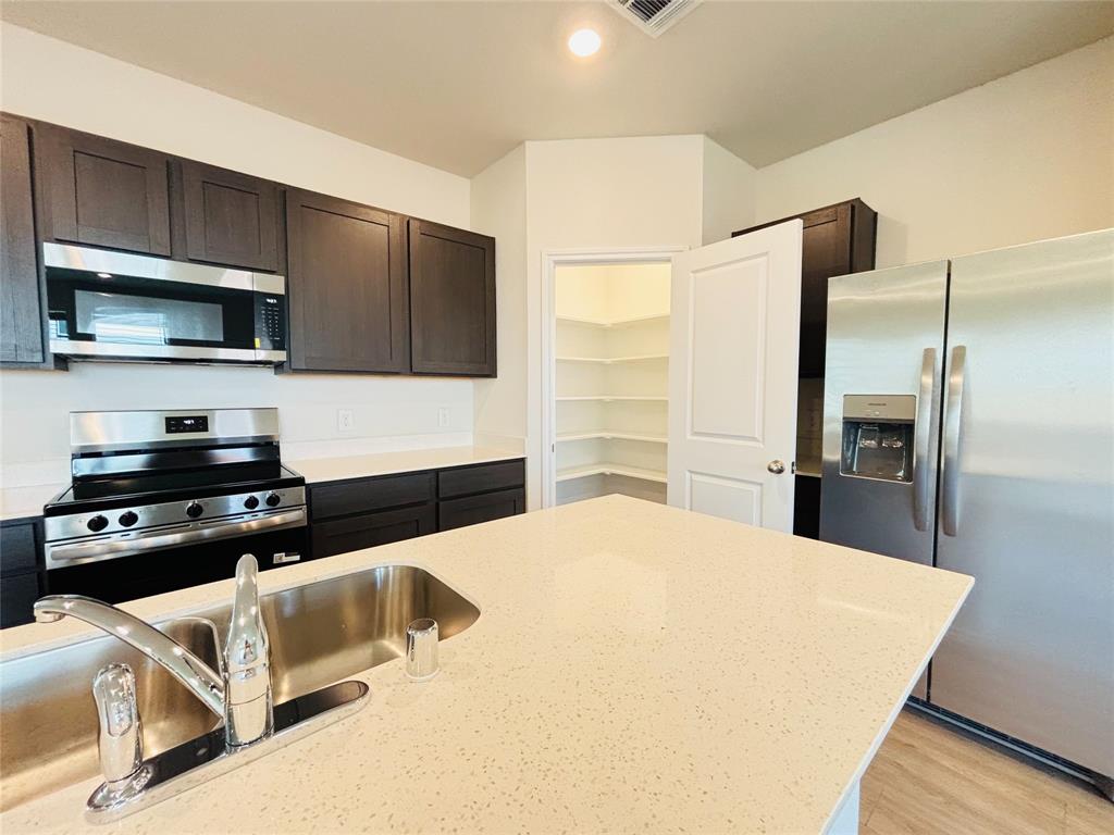 120 Redwood Road Princeton, TX 75407 - Photo 5 of 24 Modern kitchen featuring dark wood-finish cabinetry, stainless steel appliances, light-toned countertops, a stainless steel double basin sink with a chrome faucet, and a walk-in pantry with white shelving