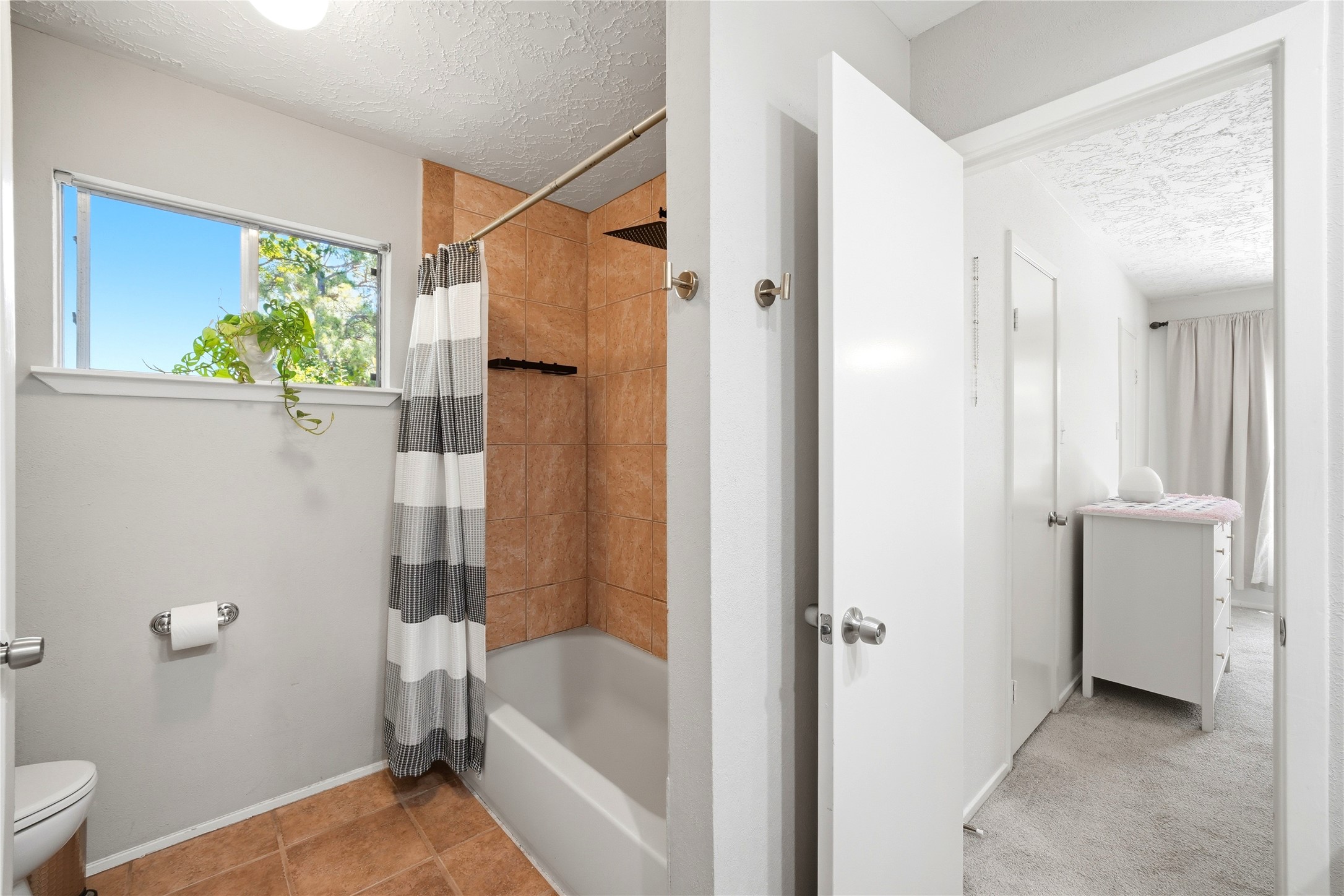 4718 Temple Bell Drive Spring, TX 77388 - Photo 35 of 50 This photo shows a bathroom with a tiled shower and bathtub, a striped shower curtain, and a small window with natural light and greenery. Adjacent is a hallway leading to a carpeted bedroom with a dresser.