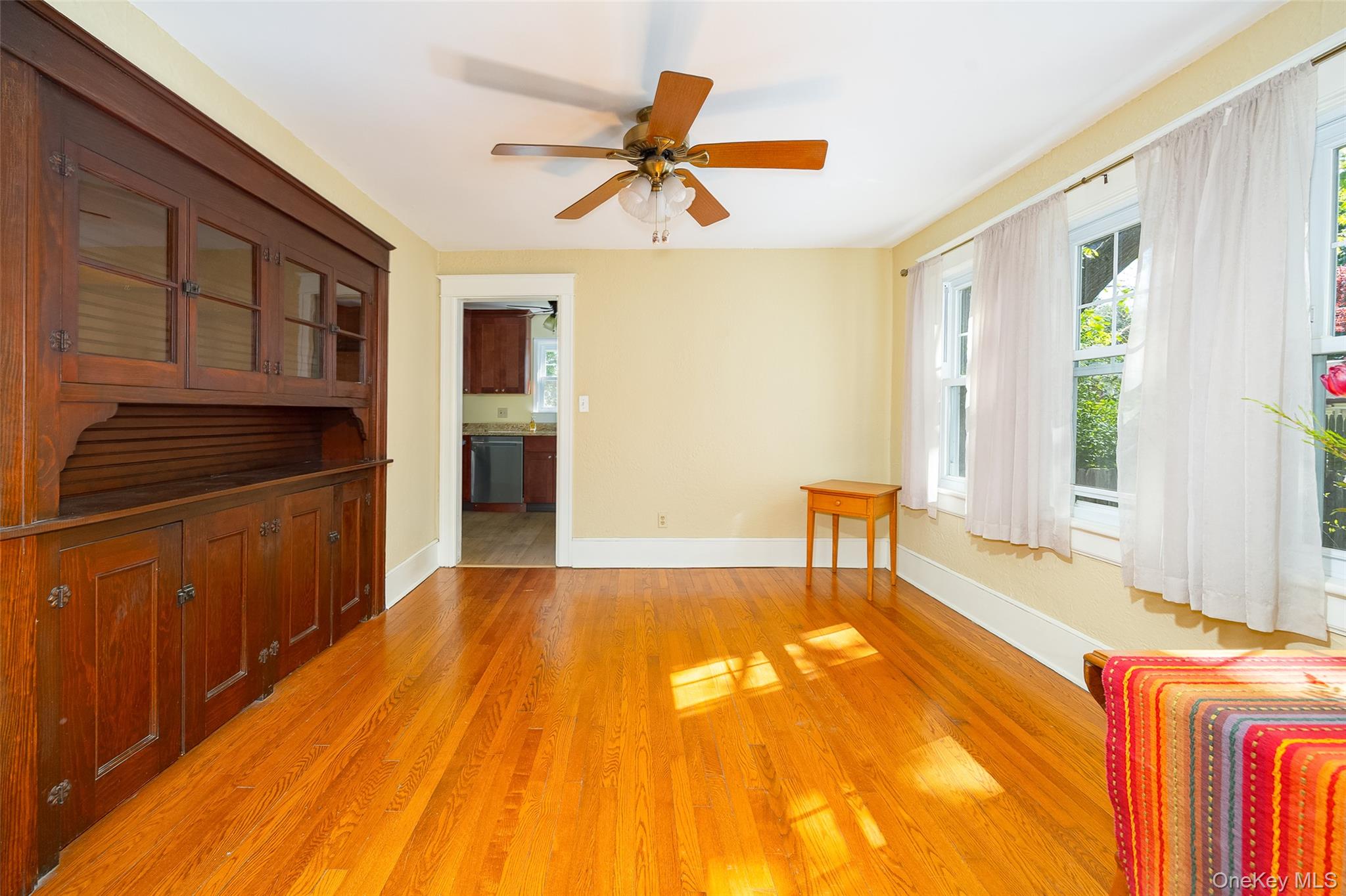 30 Marple Road Poughkeepsie, NY 12603 - Photo 12 of 26 Hardwood flooring throughout featuring a built-in wooden cabinet with glass-front uppers and lower panel doors, multiple windows with white trim, a ceiling fan with light fixture, and a doorway opening to a kitchen with wood cabinetry