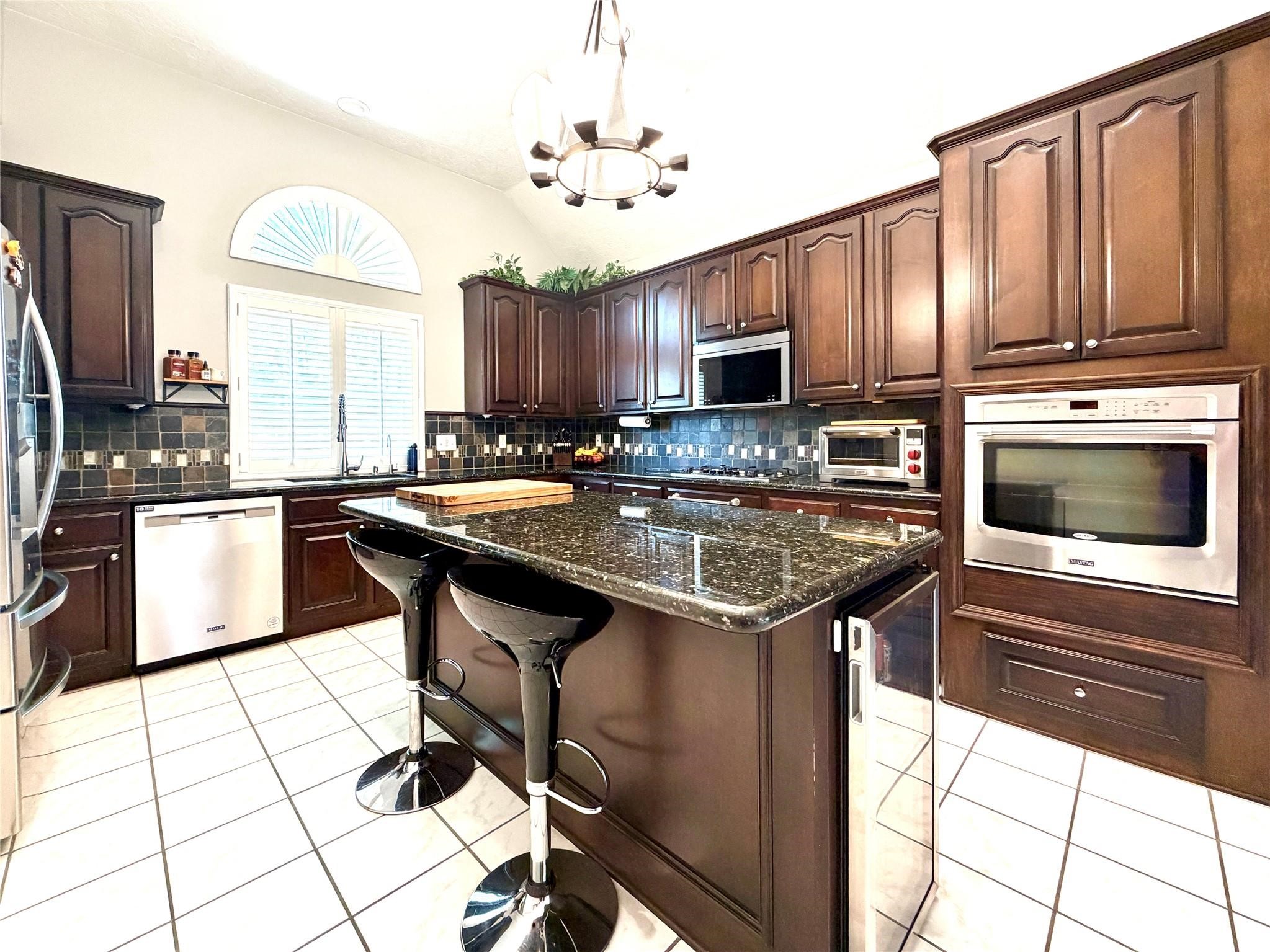 24818 Haverford Road Spring, TX 77389 - Photo 24 of 45 This kitchen features elegant dark wood cabinetry, a spacious granite island with seating, stainless steel appliances, and a stylish tile backsplash. A large arched window provides natural light, complementing the modern feel of the space.