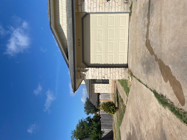 110 Verna Spur Georgetown, TX 78628 - Photo 2 of 7 A cream garage door accents the stone and shingle facade of this home, framed by a concrete driveway and mature greenery.