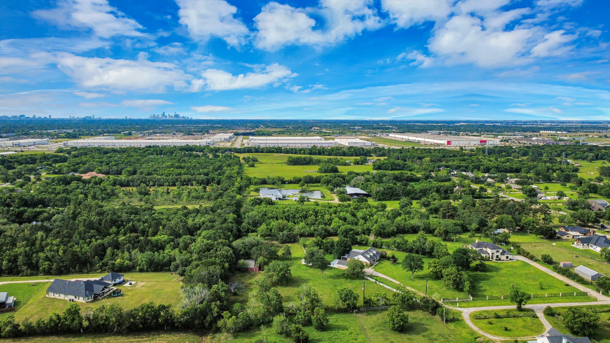 12746 Roy Road Pearland, TX 77581 - Photo 47 of 47 This aerial photo showcases a spacious, green area with scattered homes, offering a peaceful rural setting. The landscape is lush with trees and open fields, and a distant view of a city skyline is visible, suggesting convenient access to urban amenities.