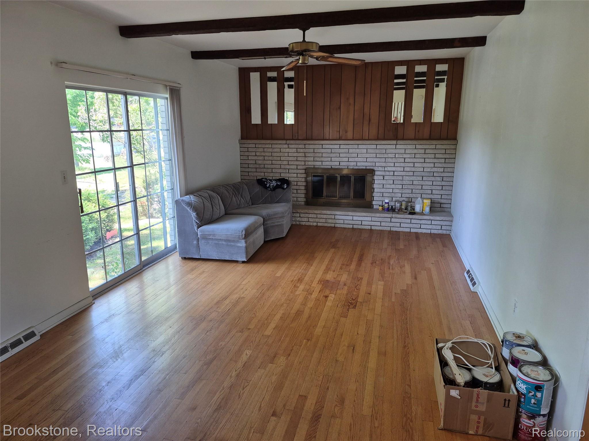 4295 Sashabaw Road Waterford, MI 48329 - Photo 18 of 37 Spacious living area featuring hardwood flooring, a large sliding glass door, a brick fireplace with a wood mantel, exposed ceiling beams, and a ceiling fan