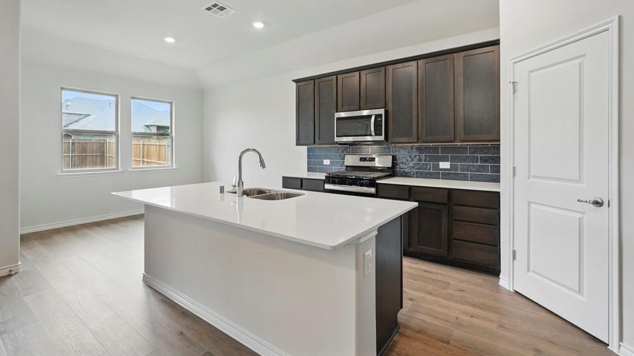 4162 Waldron Trail Forney, TX 75126 - Photo 10 of 29 Kitchen featuring a large central island with a double basin sink, dark wood cabinetry, stainless steel appliances, subway tile backsplash, and light wood-finish flooring