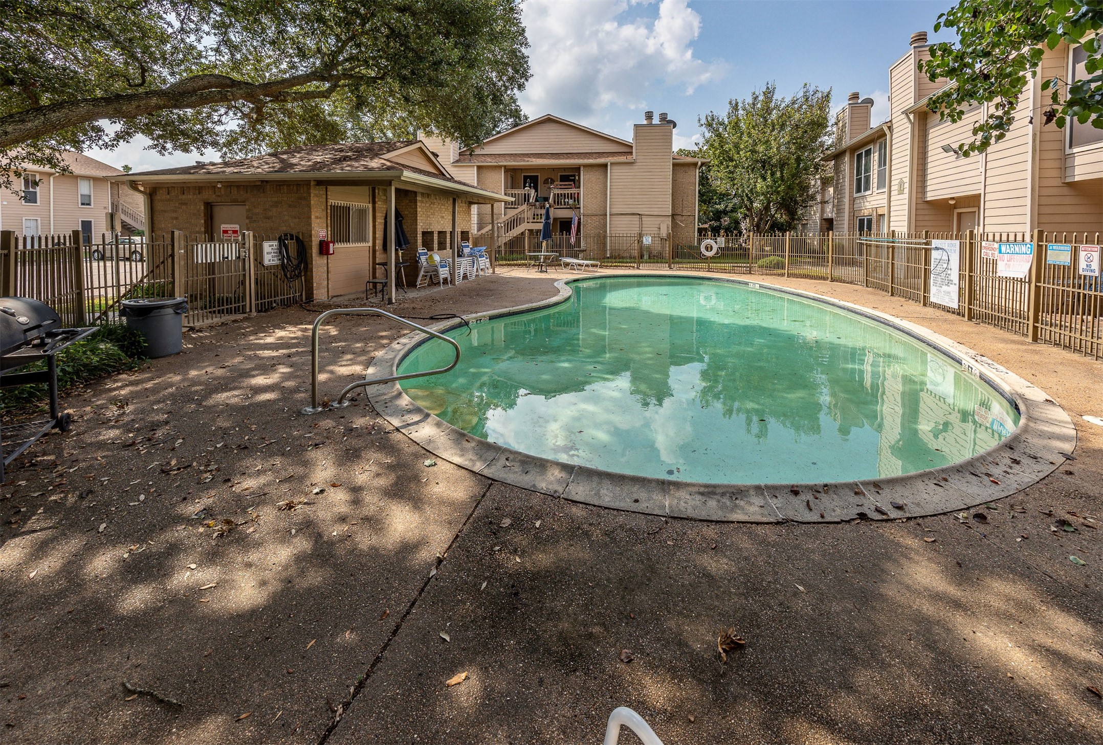 1505 Ward Road, Unit 128 Baytown, TX 77520 - Photo 20 of 21 This photo features a communal swimming pool surrounded by a paved area with seating, a small building, and nearby residential units. The area is fenced and shaded by trees, providing a relaxing outdoor space.
