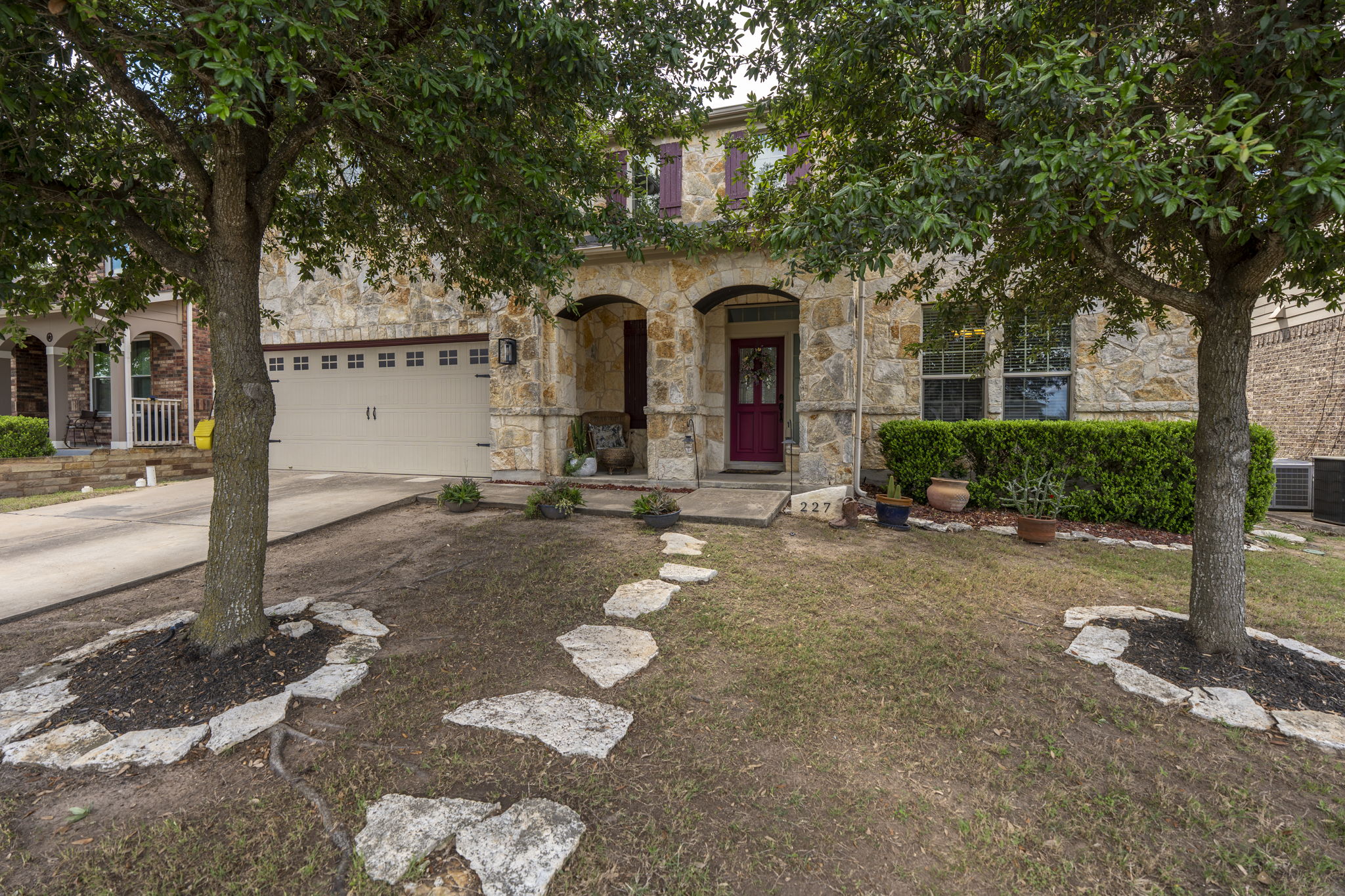 227 Sandstone Trail Buda, TX 78610 - Photo 33 of 33 Stone facade home featuring a two-car garage, red entry door, covered front porch, and mature trees