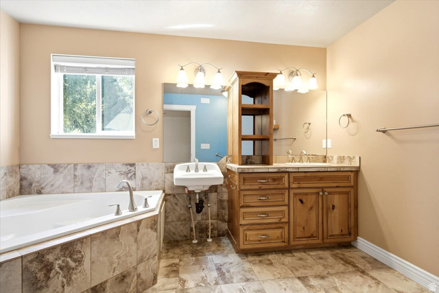 1089 West 2325th Street North Layton, UT 84041 - Photo 21 of 40 Bathroom featuring a corner soaking tub with tile surround, a double vanity with wood cabinetry and a stone-look countertop, a wall-mounted sink, and tile flooring