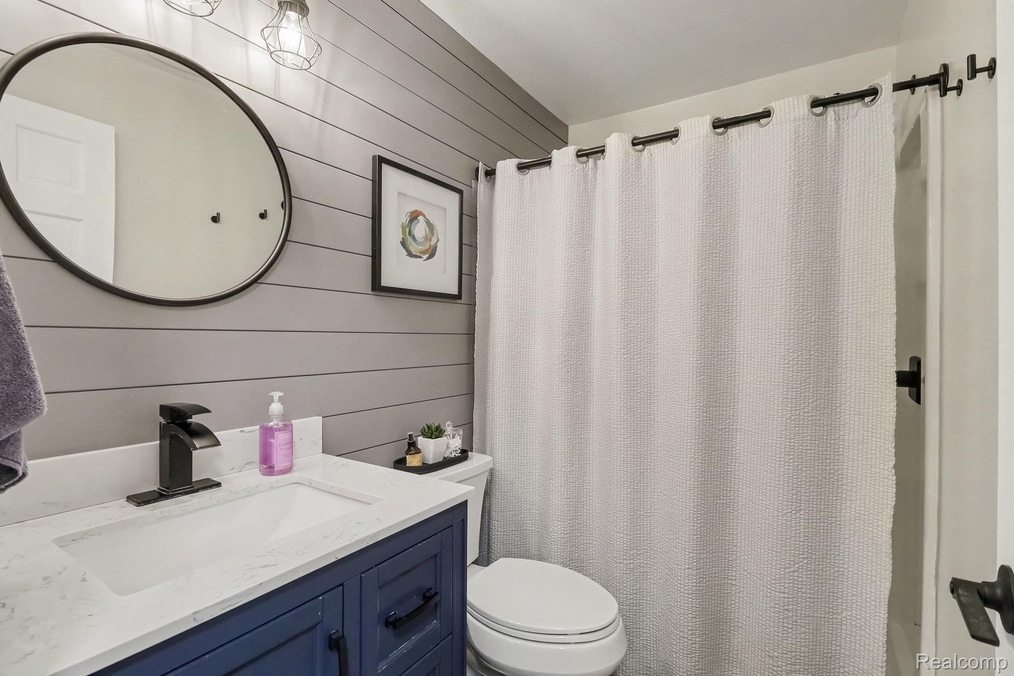 24404 Jamestowne Road Novi, MI 48375 - Photo 15 of 26 Vanity with a white countertop and blue cabinetry, a toilet, a round mirror, and a shower with a white curtain