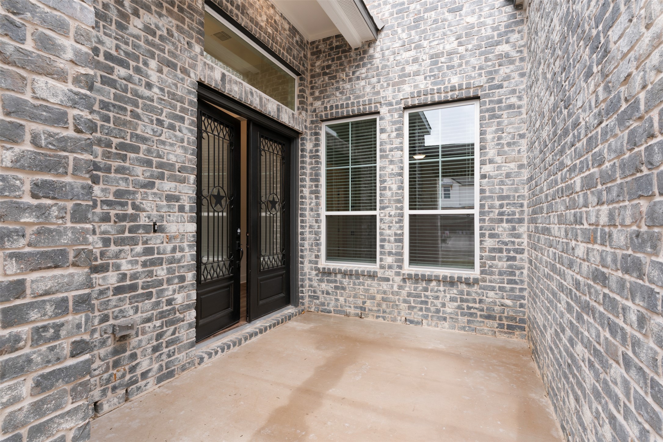 2312 Tobiano Trace Georgetown, TX 78633 - Photo 4 of 33 Covered entryway featuring a double-door entrance with wrought iron inserts, an overhead transom window, and two vertical windows with integrated blinds, all set against a gray brick facade and concrete flooring
