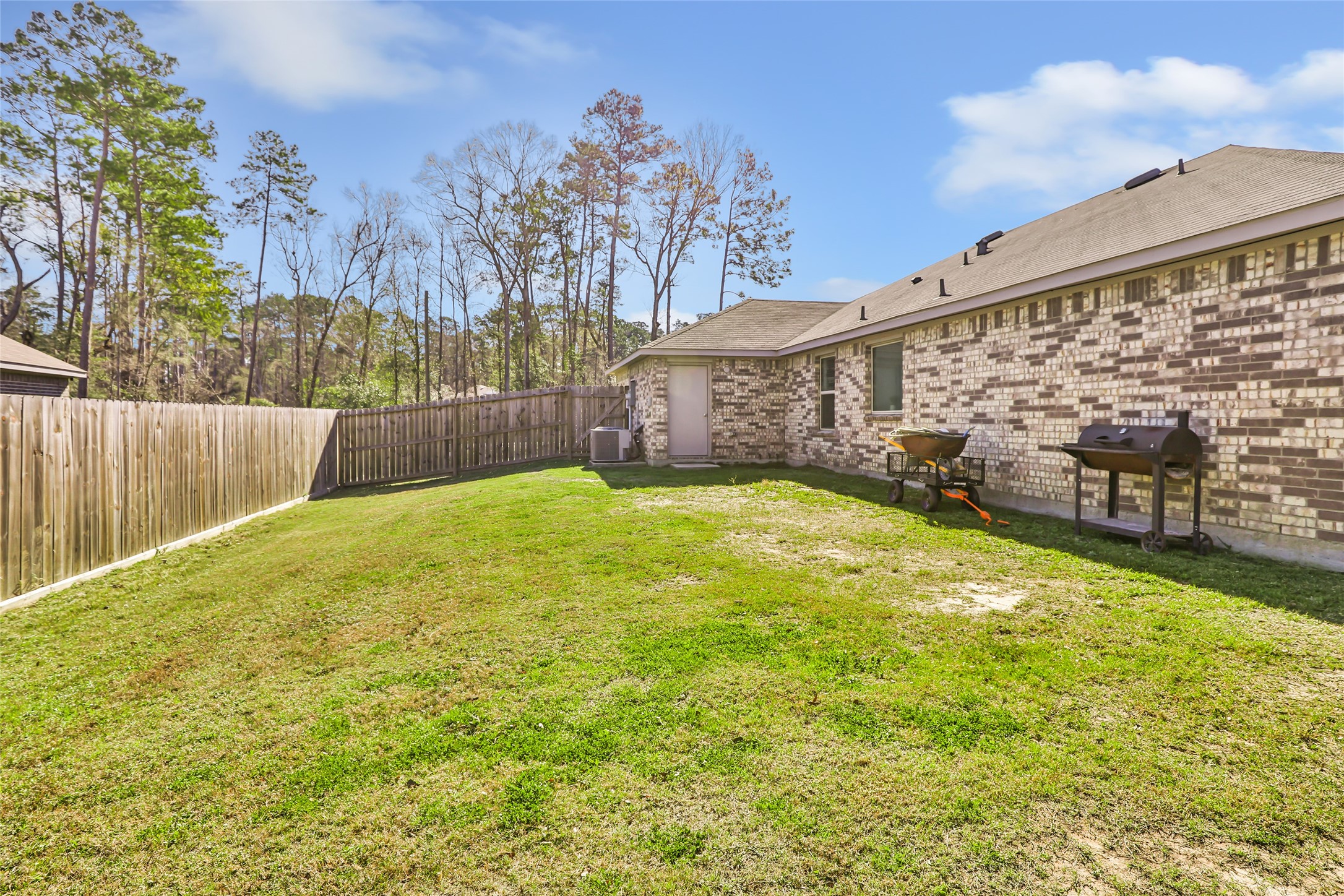 811 Republic Road New Caney, TX 77357 - Photo 12 of 37 This photo shows a spacious backyard with a well-maintained lawn, bordered by a wooden fence for privacy. There's a brick exterior house with a grill and a wheelbarrow, ideal for outdoor activities and entertaining. Tall trees provide a scenic backdrop.