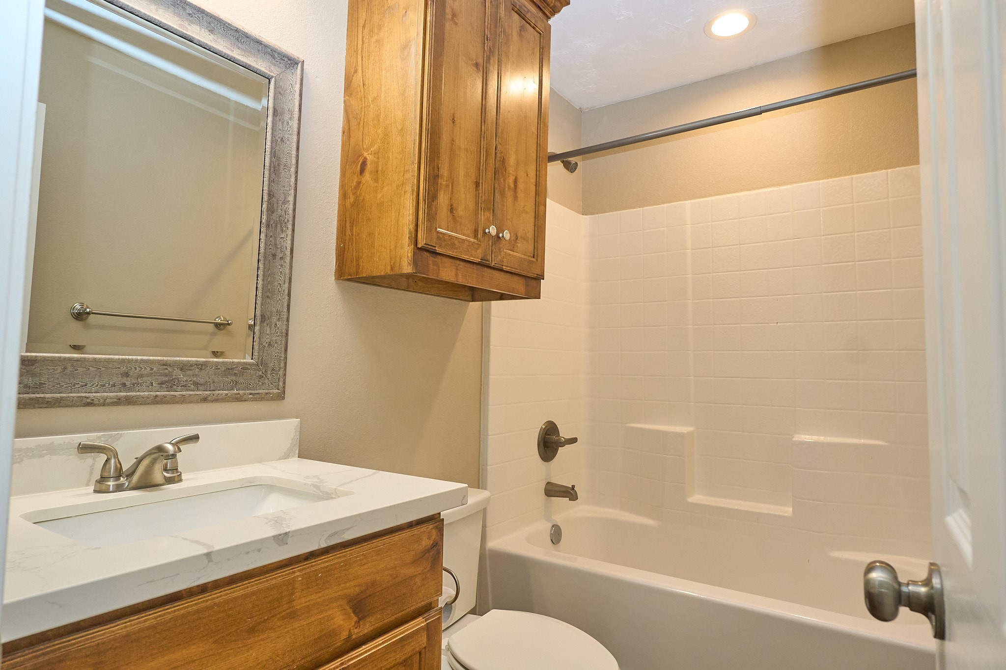 1807 Nuches Lane Bryan, TX 77803 - Photo 21 of 27 This bathroom features a modern design with a white bathtub and shower combination, light-colored tiles, and a wood vanity with a marble countertop. A rustic framed mirror and overhead cabinet provide additional storage space.