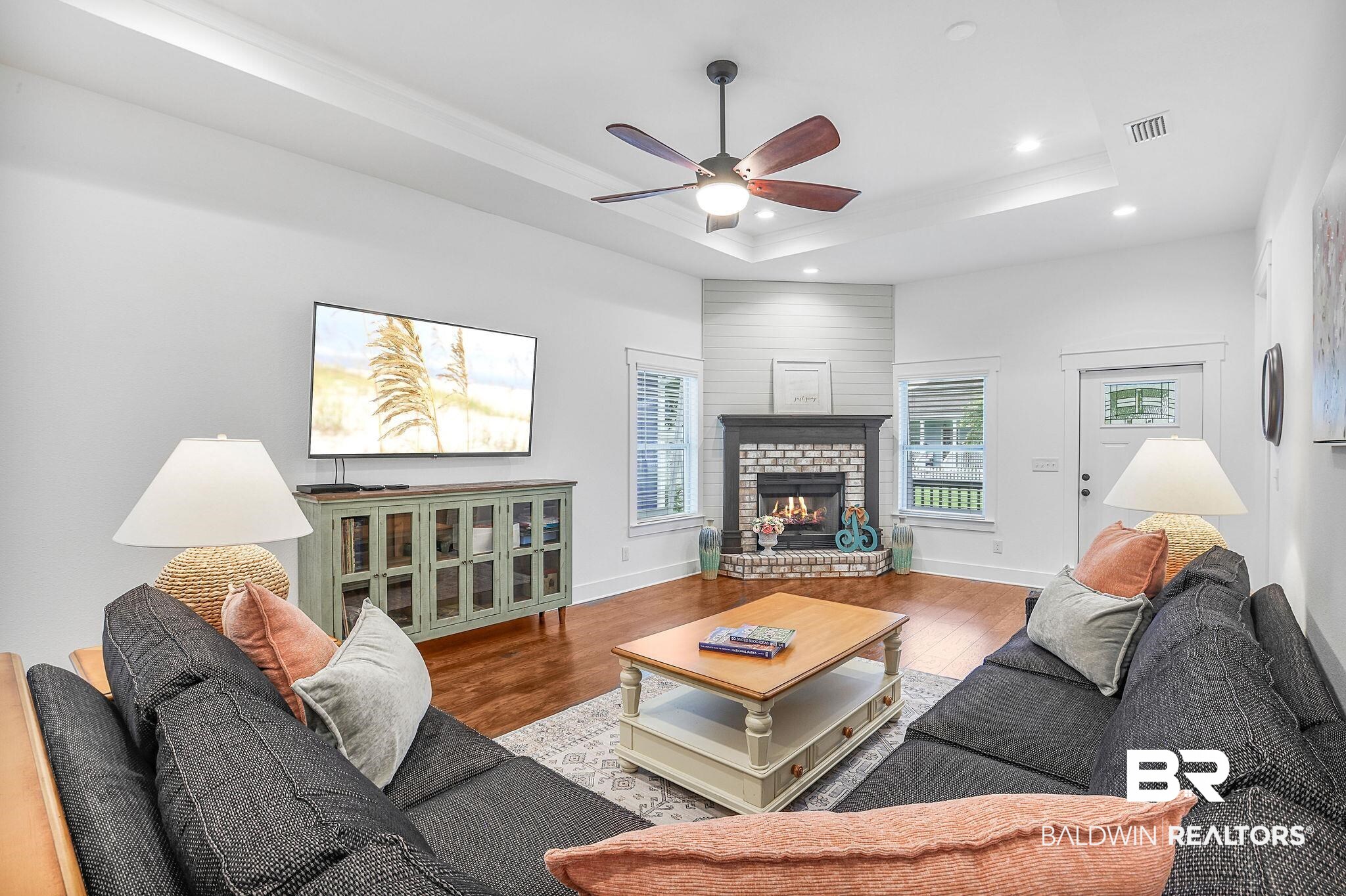 Living area featuring ceiling fan, wood finished floors, a tray ceiling, recessed lighting, and a brick fireplace