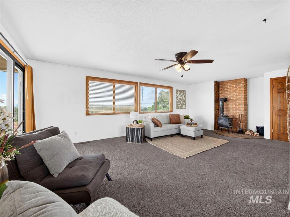 8989 Hillview Road Emmett, ID 83617 - Photo 10 of 50 Living area with neutral carpeting, a brick-faced corner hearth featuring a wood-burning stove, and multiple windows with wood-frame blinds