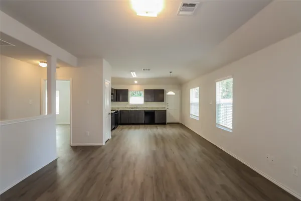 a view of an empty room and a kitchen with wooden floor and windows