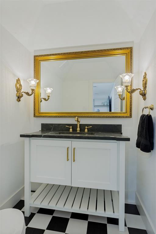 4316 Springbranch Drive Benbrook, TX 76116 - Photo 9 of 39 Vanity featuring a black and white checkerboard floor, white shaker-style cabinetry with gold hardware, a black stone countertop, and a gold-framed mirror flanked by ornate sconces