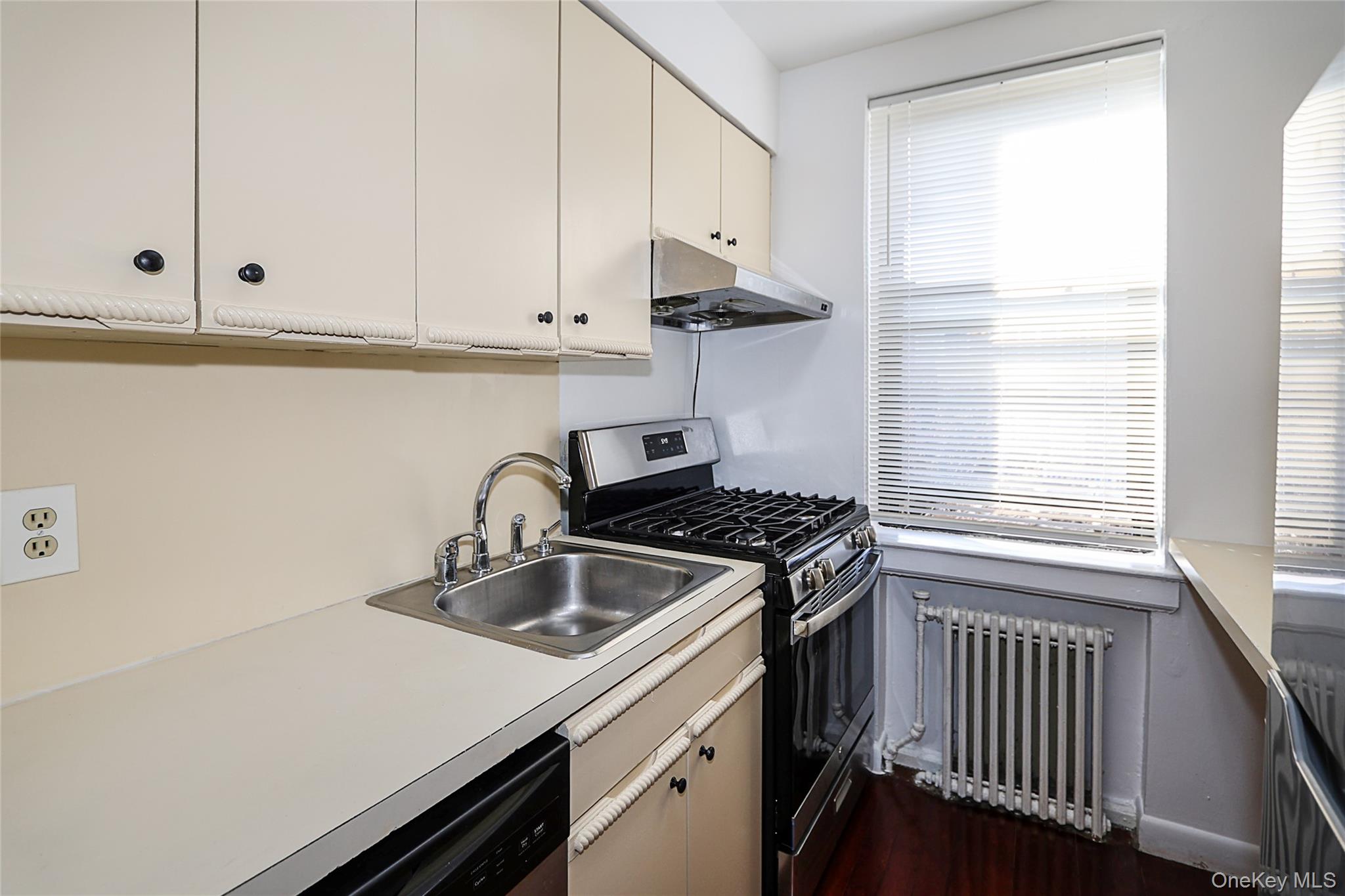47-22 215th Place, Unit 2C Queens, NY 11361 - Photo 14 of 26 Kitchen with stainless steel appliances, radiator heating unit, light countertops, under cabinet range hood, and dark wood-style floors