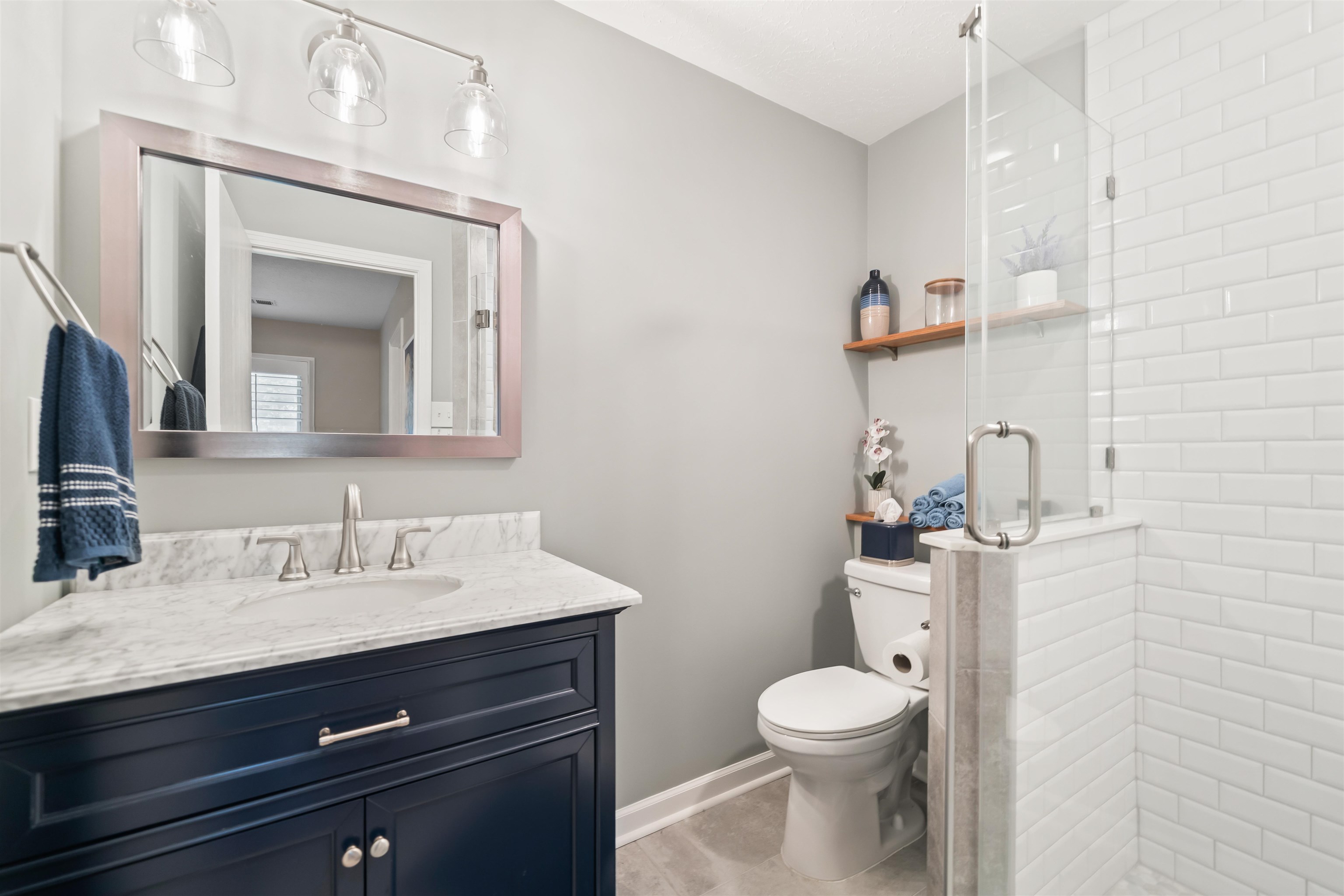 6943 Fords Station Road, Unit 64 Germantown, TN 38138 - Photo 25 of 30 Bathroom featuring a dark blue vanity with a white marble-finish countertop, a frameless shower with subway tile walls, and a recessed toilet area with wood shelving
