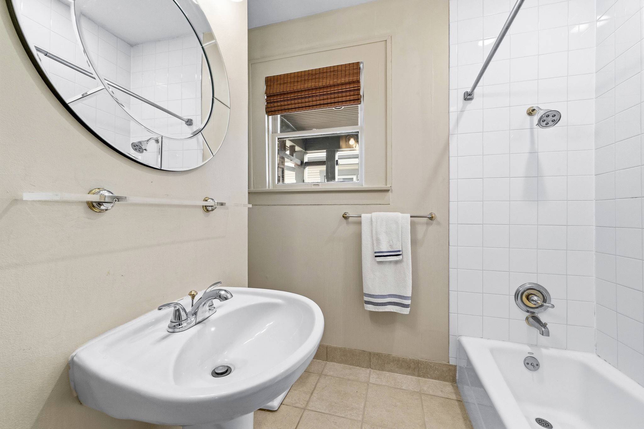 1521 Kipling Street Houston, TX 77006 - Photo 29 of 35 Compact bathroom featuring a white pedestal sink, a circular mirror, and a tiled shower-tub combo. Light beige walls and a window with a bamboo shade add a warm touch.