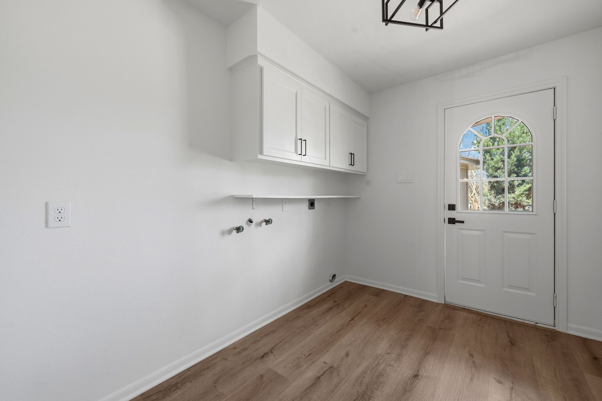 210 Ridgemont Drive Alvin, TX 77511 - Photo 10 of 27 This photo features a bright utility room with white walls and wood flooring. It includes overhead cabinets for storage, a door with a decorative window, and hookups for laundry appliances.