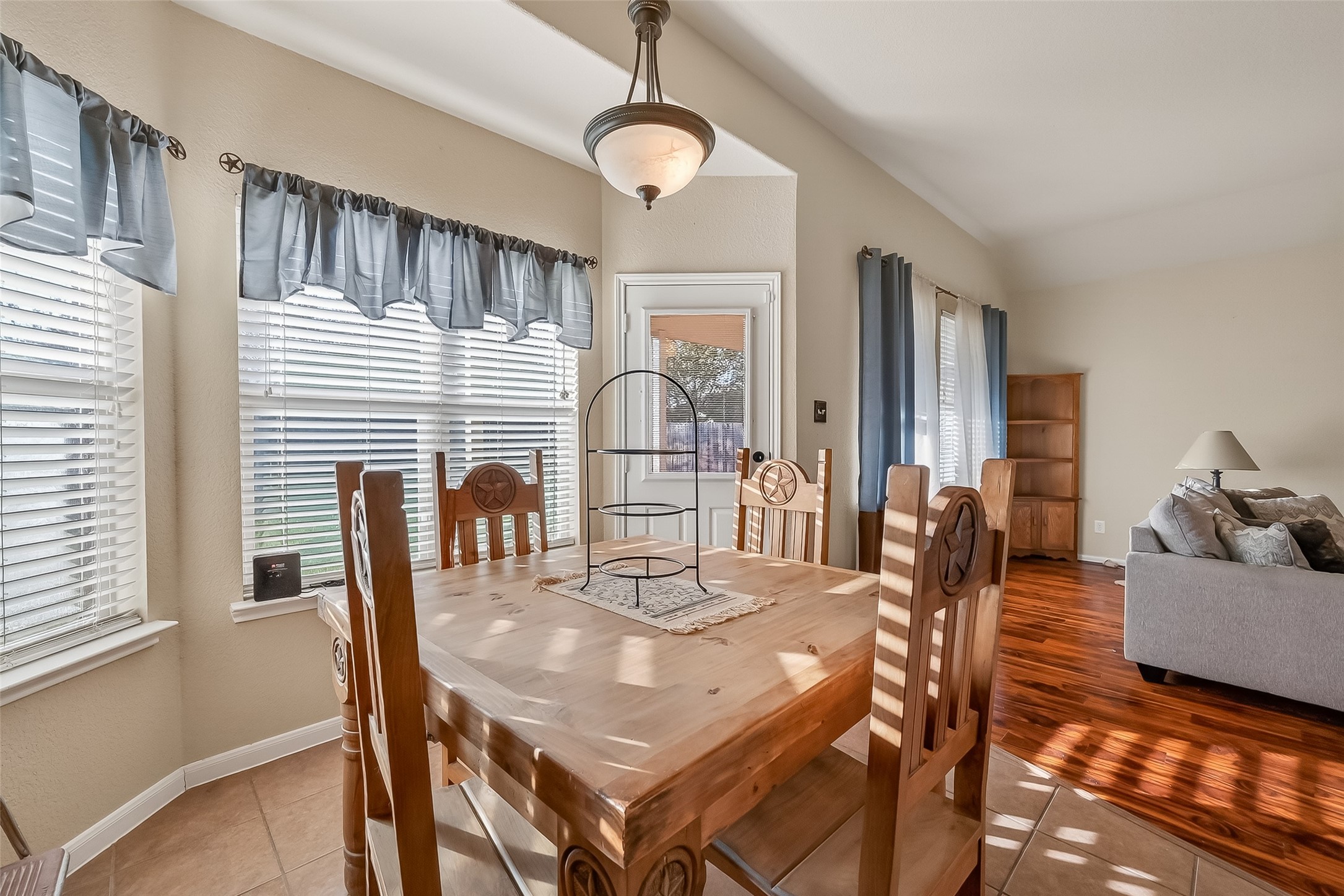 This bright dining area features large windows with blue curtains, a wooden table set, and tile flooring. It opens into a cozy living space with hardwood floors.