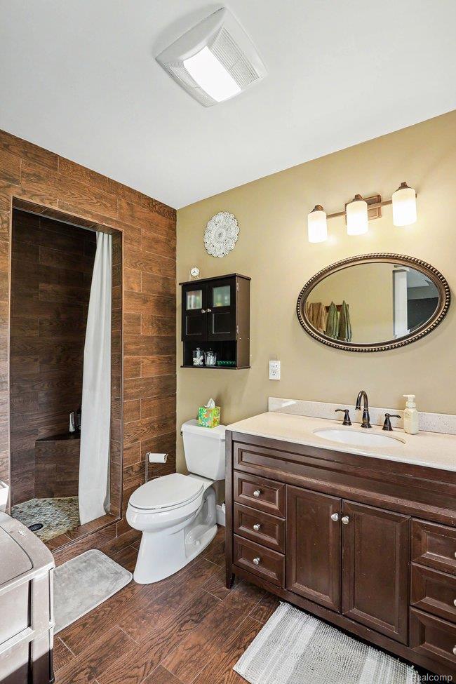 5793 Maple Grove Road Silverwood, MI 48760 - Photo 13 of 33 Bathroom featuring wood-finish floor tiling, a walk-in shower with tiled walls and a built-in bench, a dark wood vanity with a light countertop and undermount sink, an oval mirror, and a three-light fixture