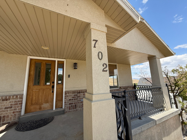702 Elk Meadow Loop Tooele, UT 84074 - Photo 2 of 9 Covered porch entry featuring a wood-panel door with glass inserts, textured stucco siding, brick detailing, and decorative wrought iron railing