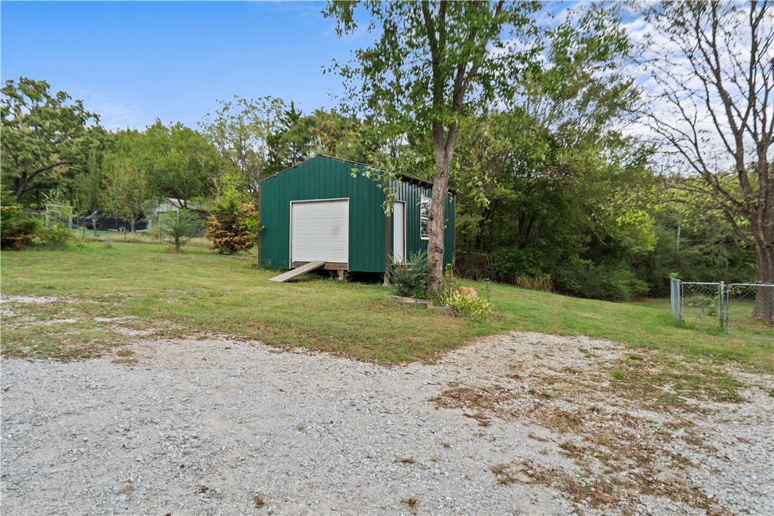 230 South Stone Bridge Road Fayetteville, AR 72701 - Photo 4 of 34 Gravel driveway, expansive grassy area, metal outbuilding with a roll-up door, mature trees, and chain link fencing