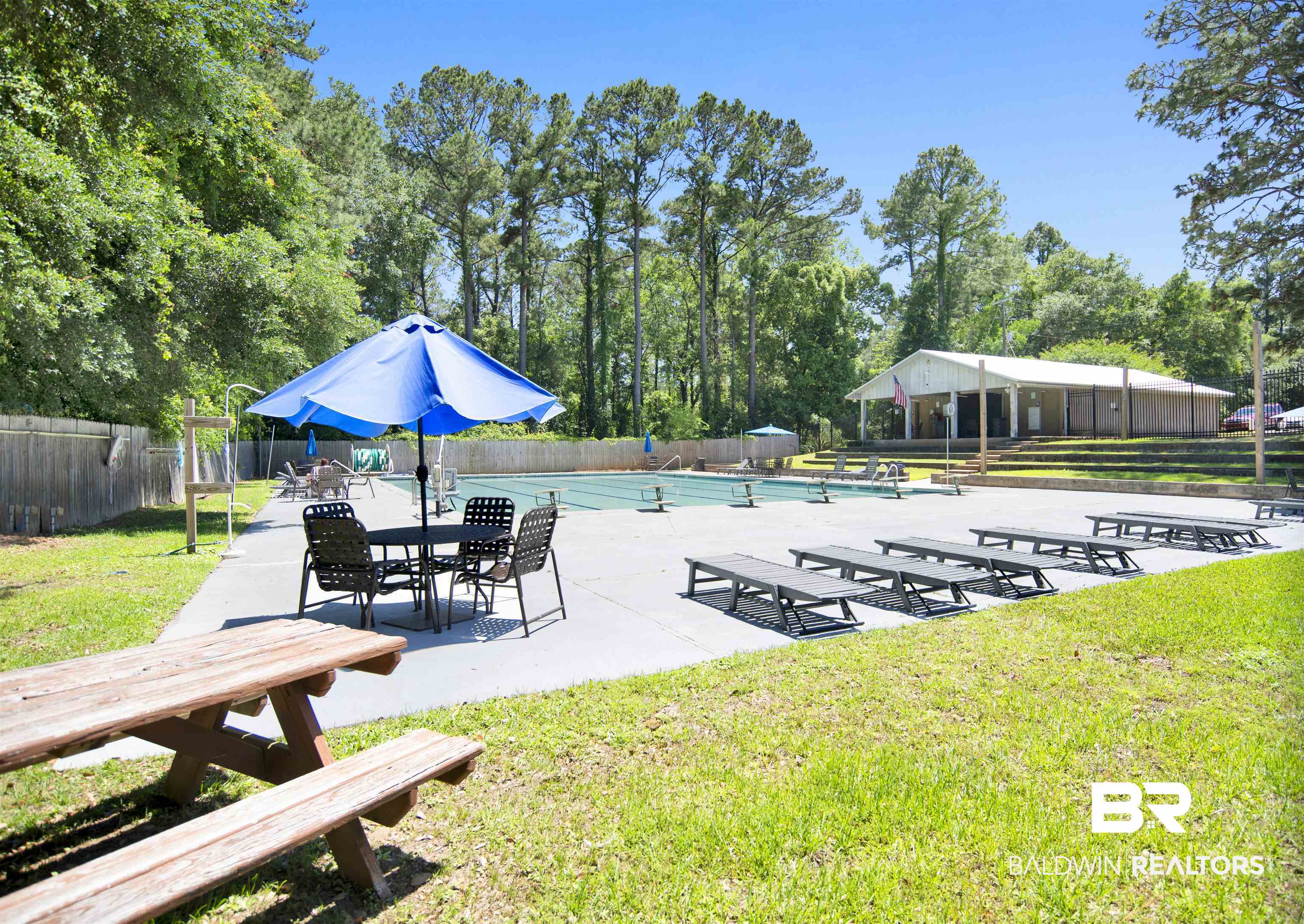 107 Virginia Circle Daphne, AL 36526 - Photo 41 of 47 Outdoor swimming pool with a concrete surround, surrounded by mature trees, a wooden fence, and a distant building with a covered patio
