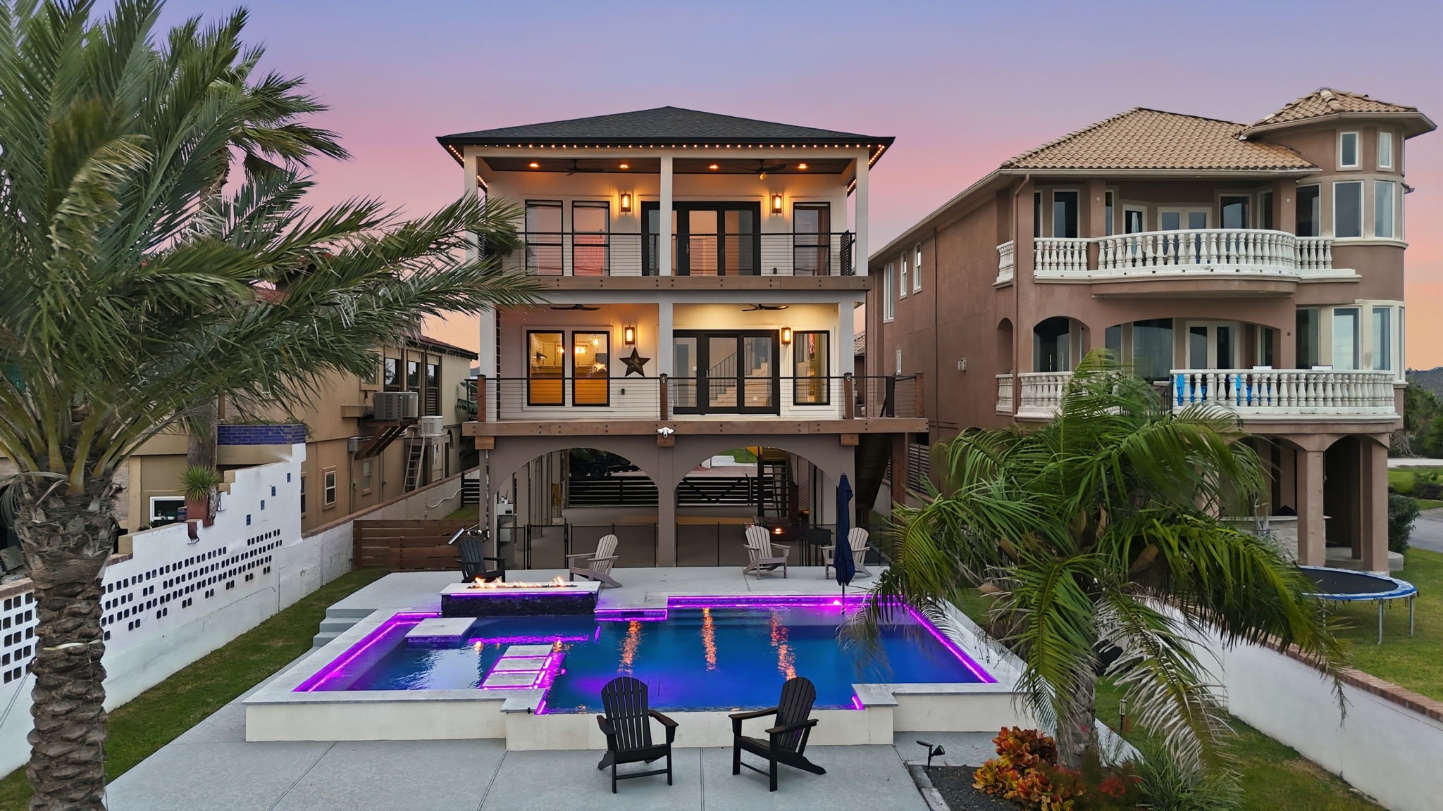 Sunset frames this luxurious bayfront retreat, showcasing a resort-style pool with LED accents, fire feature, pier, and multiple lounging areas on an oversized waterfront lot.