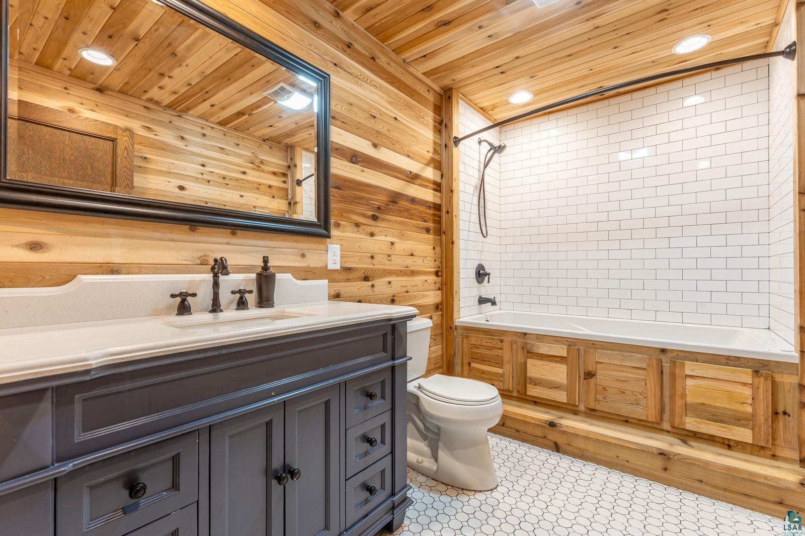 9734 Alango Road Angora, MN 55703 - Photo 11 of 41 Bathroom featuring extensive wood paneling, a dark grey vanity with a light countertop, a framed mirror, a tub/shower combination with white subway tile surround, and white hexagonal floor tiling