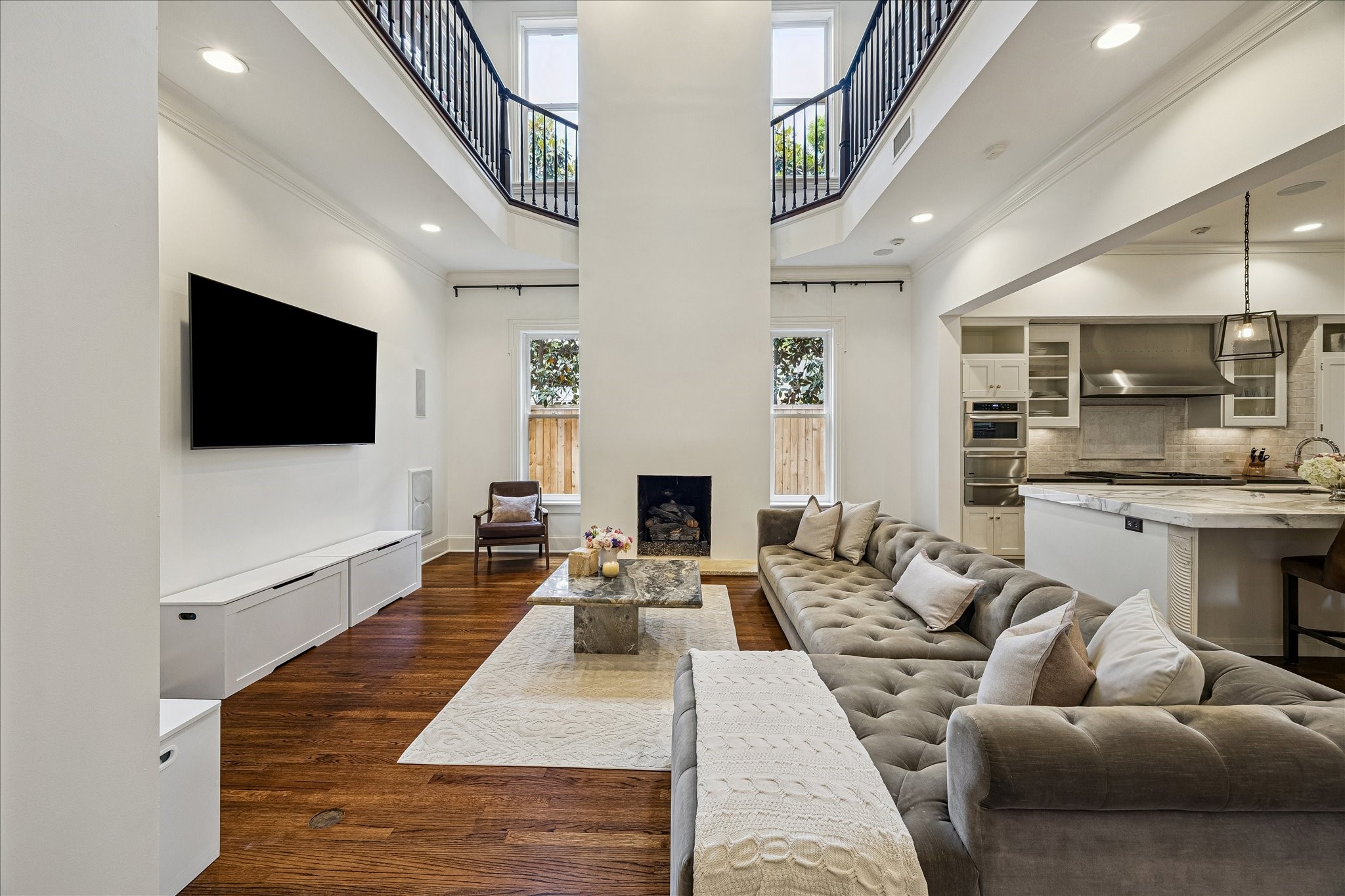 2368 Timber Lane Houston, TX 77027 - Photo 16 of 46 The living room strikes a rare balance between grandeur and comfort, anchored by soaring ceilings, rich hardwood floors, and an airy open-concept design filled with natural light from surrounding windows above and below. Dramatic ceiling heights draw the eye upward into the second-floor library gallery overlooking the space, creating a striking architectural presence while maintaining an inviting atmosphere.