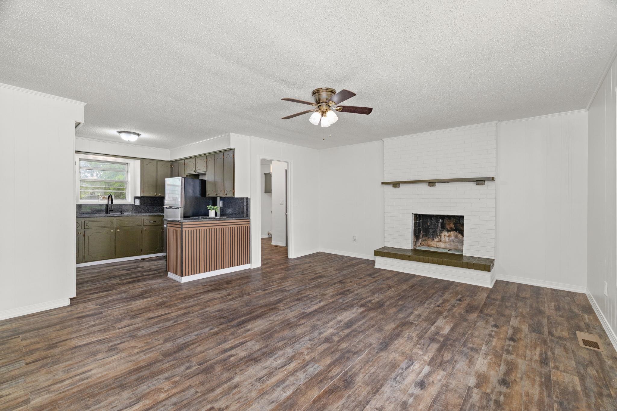 217 Red Bay Road Golden, MS 38847 - Photo 6 of 33 Open-concept living space featuring wood-finish flooring, a white brick fireplace with a wood mantel, a ceiling fan, and a kitchen area with olive green cabinetry, dark countertops, and a black sink with a gooseneck faucet