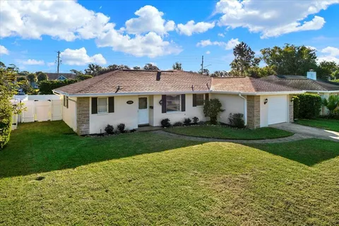 a view of a white house in front of a big yard with plants and large trees