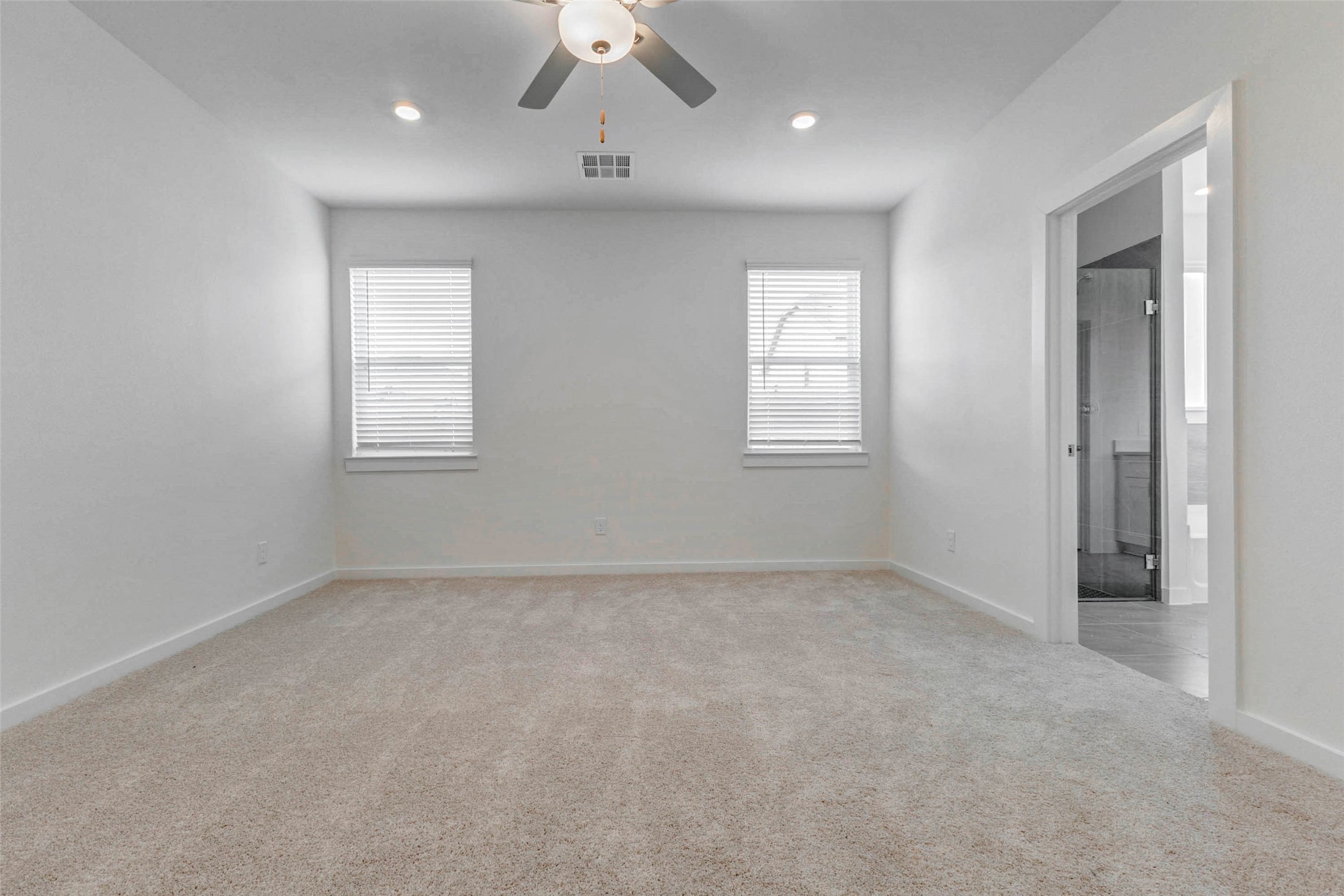 6471 Escondido Drive Rosharon, TX 77583 - Photo 18 of 33 Spacious primary bedroom with soft carpet, neutral walls, recessed lighting, ceiling fan, and two windows for natural light. The private en-suite bath entrance adds convenience, while the open wall space and calm color palette make the room easy to personalize as a peaceful retreat.