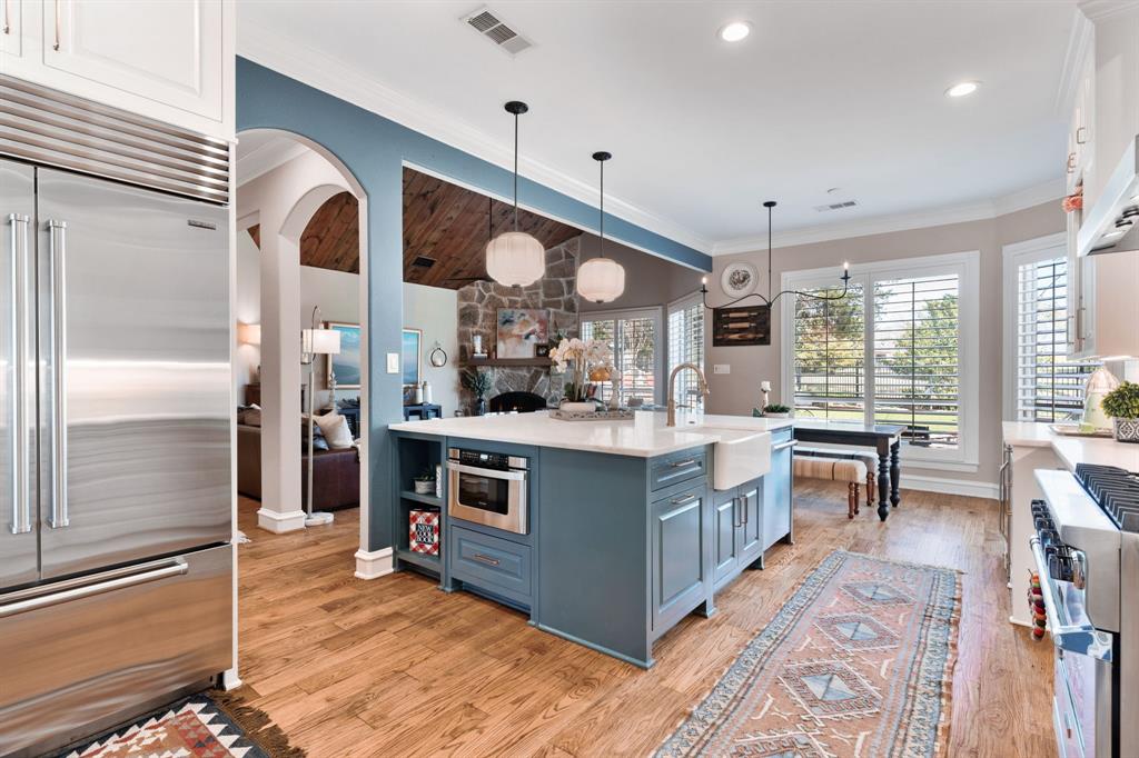 2404 Racheal Drive Heath, TX 75032 - Photo 13 of 40 Hardwood floors complement the honed marble countertops that extend seamlessly into the backsplash, while a massive eat-in island—accented with rich blue cabinetry and open shelving—offers abundant storage and gathering space. A white DXV farmhouse sink is complemented by a soft gold pull-down faucet, merging style with everyday functionality, while two distinctive Anthropologie pendant lights grace the island